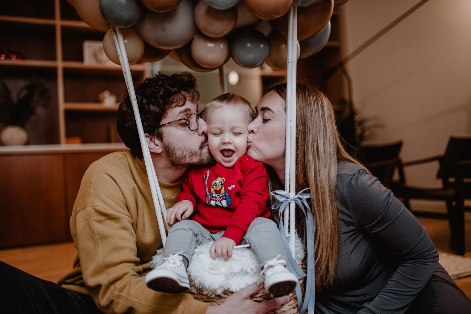 Family sharing loving kisses with baby under grey balloon decorations at indoor celebration.