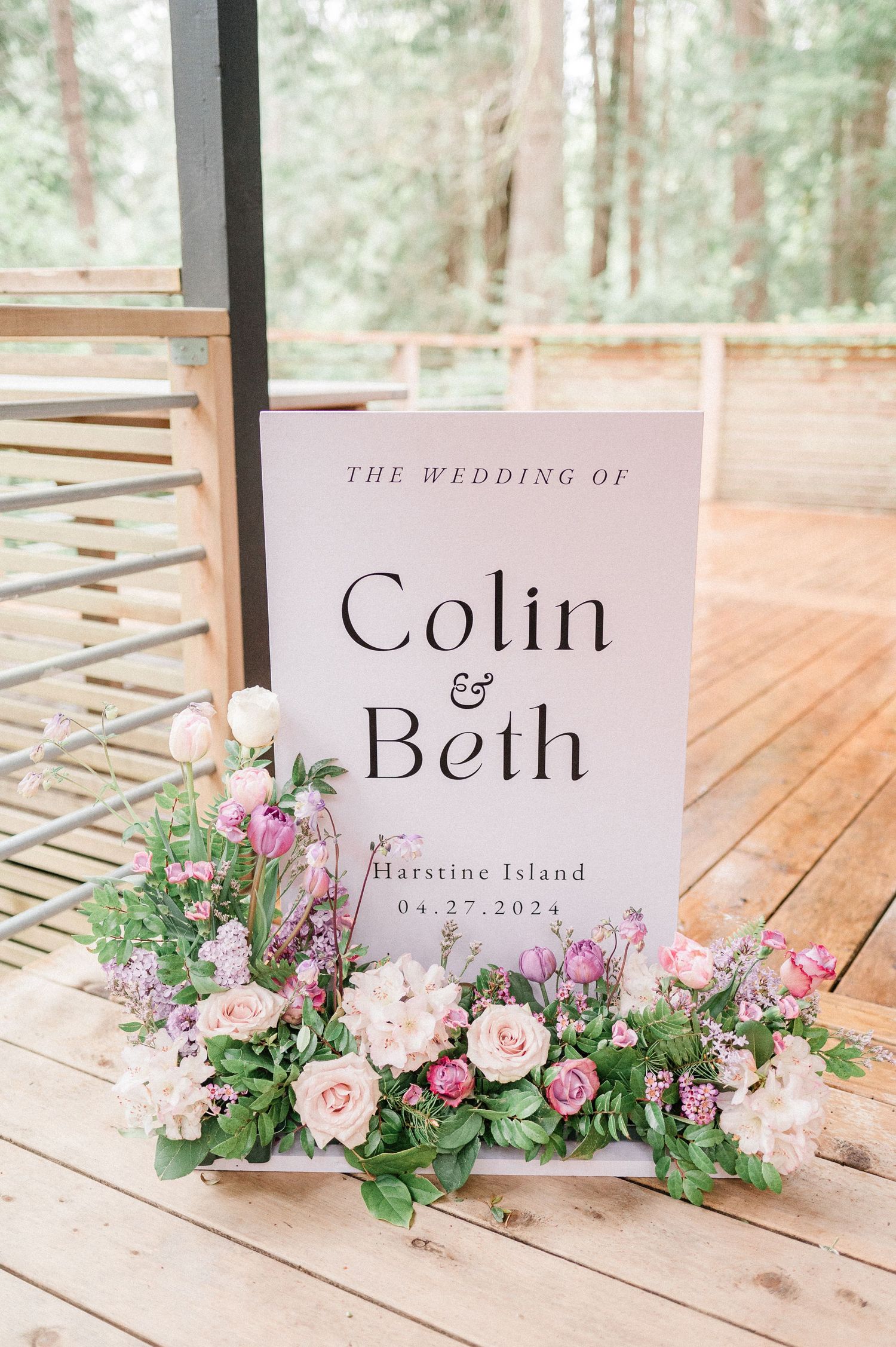 Elegant wedding signage with floral arrangements featuring pink roses and greenery displayed on wooden deck