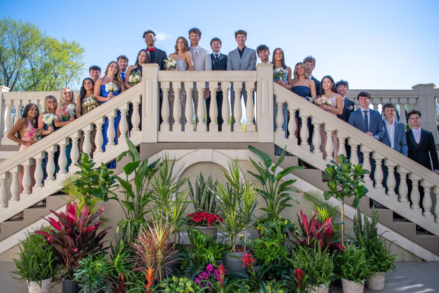 Large group poses on ornate white stone staircase with tropical plants and flowers.