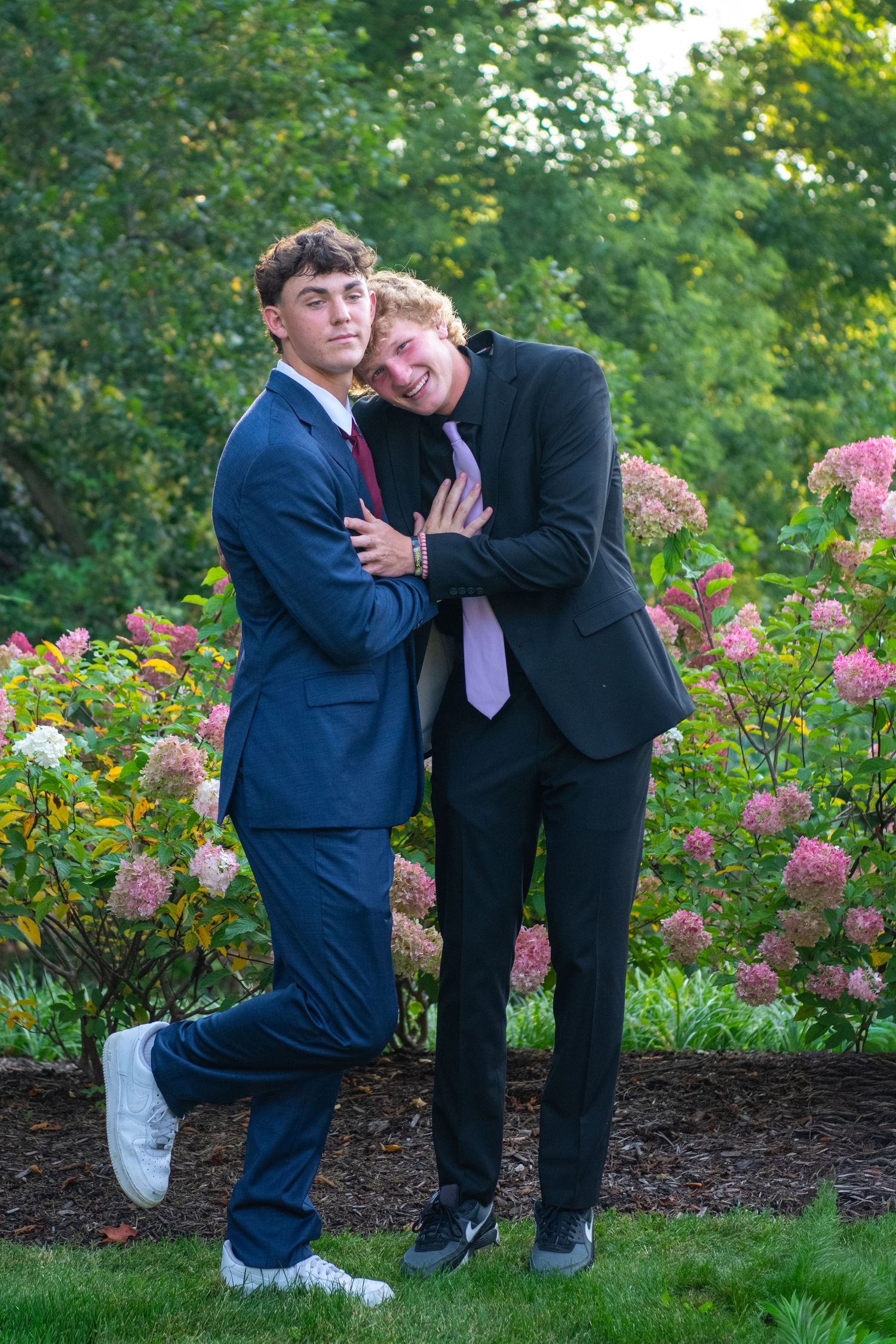 Two people in formal attire share a playful moment in a garden with pink flowers blooming in the background.
