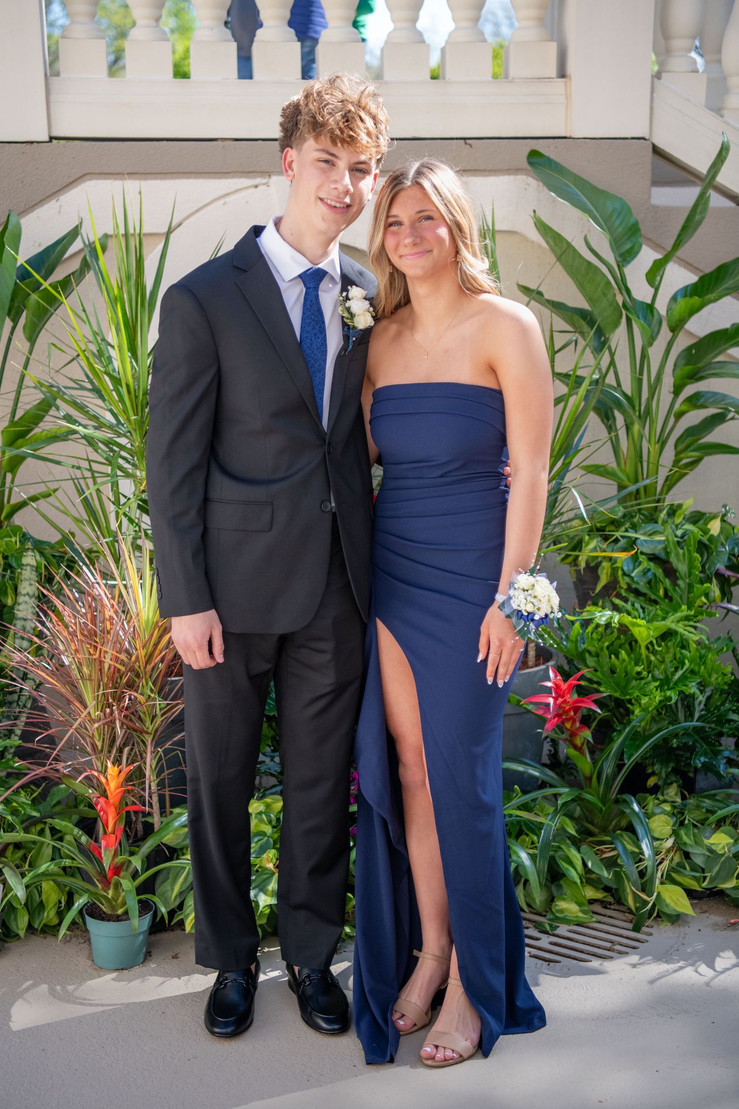 Formal couple in navy and black attire pose together against white railing backdrop.
