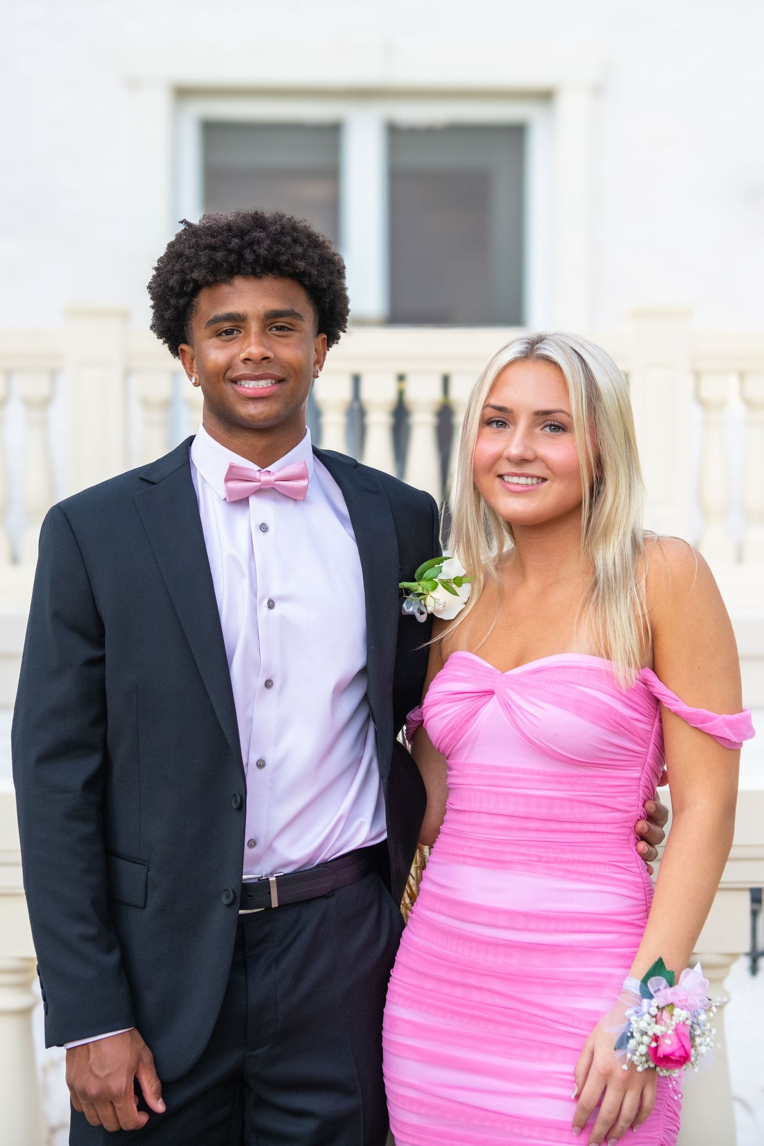 A couple poses together at a formal event wearing a black tuxedo and pink off-shoulder dress.
