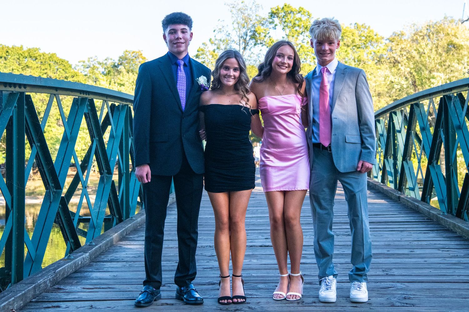Four young people dressed in formal attire stand together smiling on a green metal bridge on a sunny day.