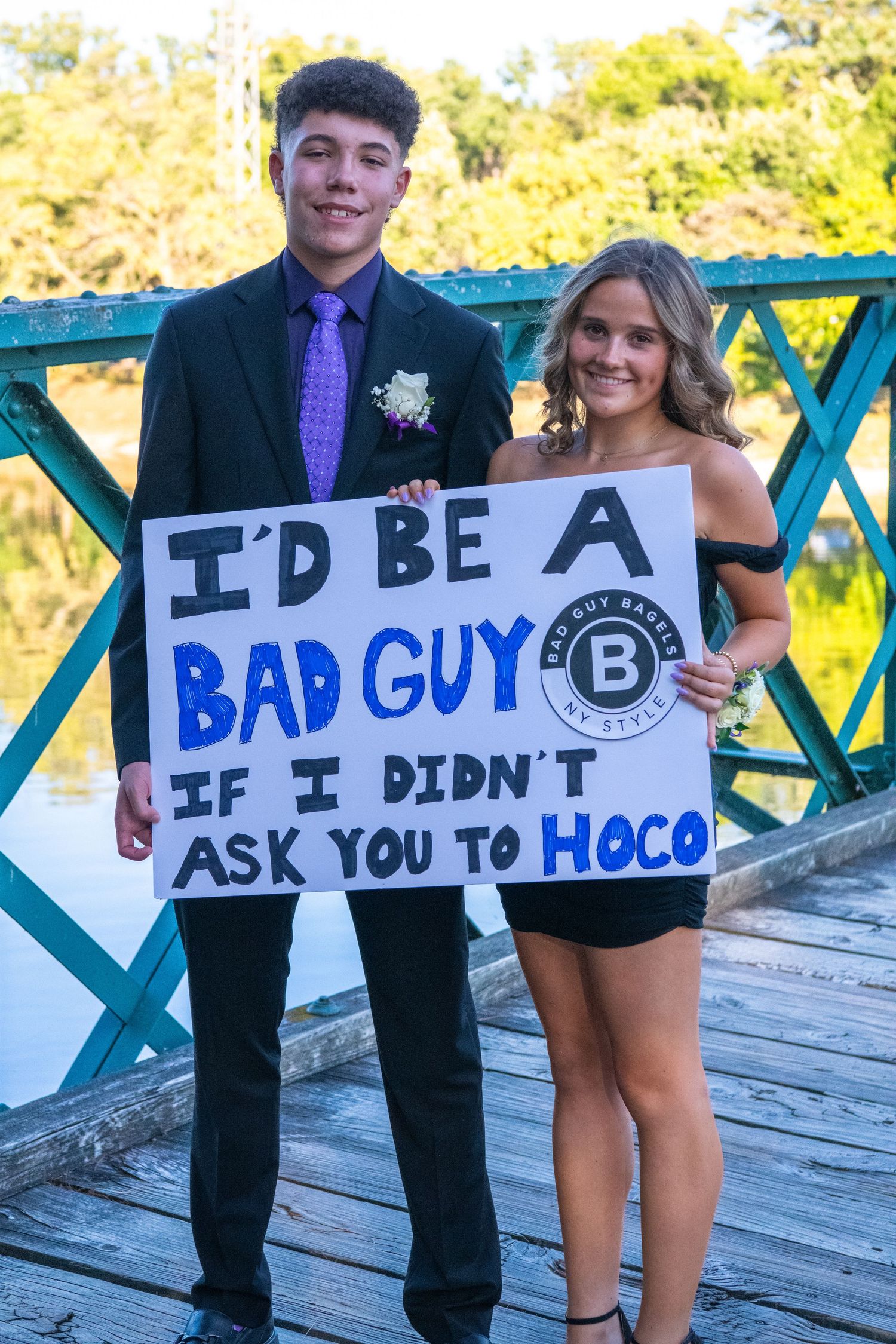 A creative prom proposal sign held up piece by piece on a wooden deck with teal railings in outdoor lighting.