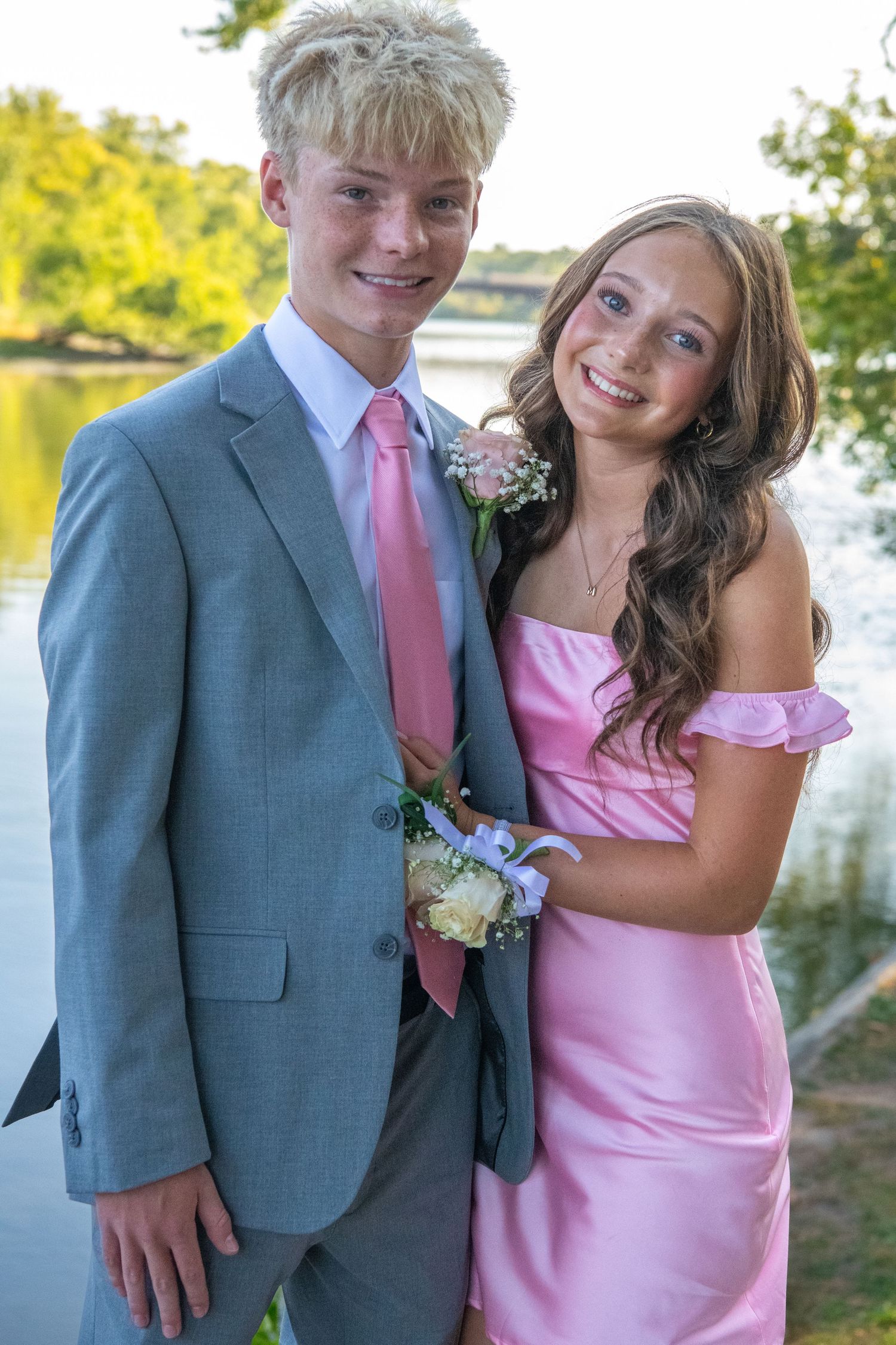 A couple in formal attire poses outdoors, with the person wearing a gray suit and pink tie next to someone in a pink dress.
