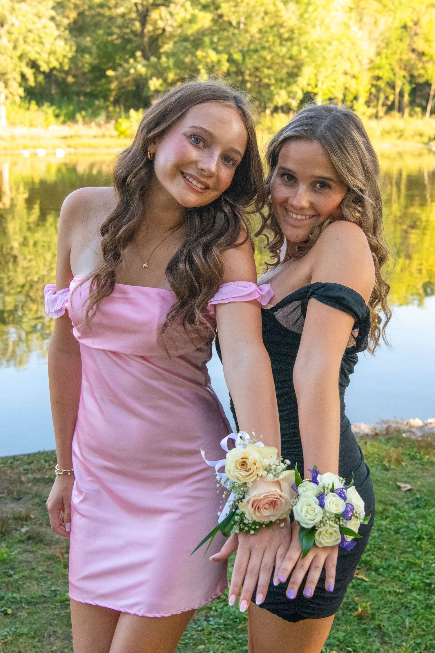 Two friends in pink and black dresses show off their matching corsages by a lake during golden hour.