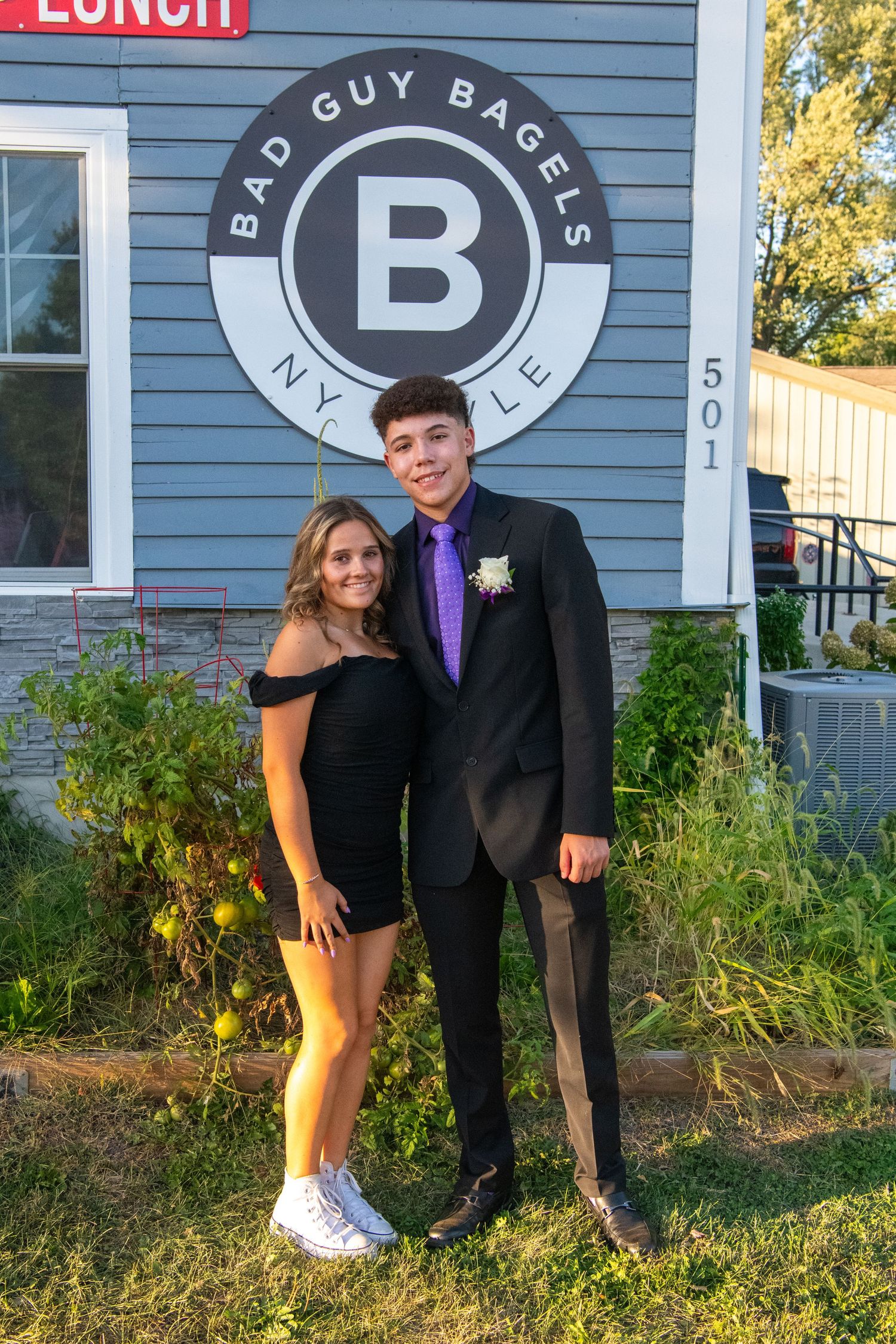 A couple poses together outdoors in formal attire near a blue building and garden at sunset.