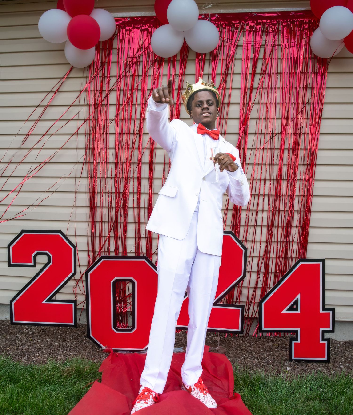 Graduate in white suit poses against red and white decorations with class year numbers.