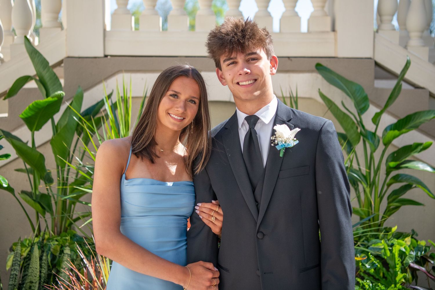 Wedding party members pose against a white balustrade wall with tropical plants in the background.