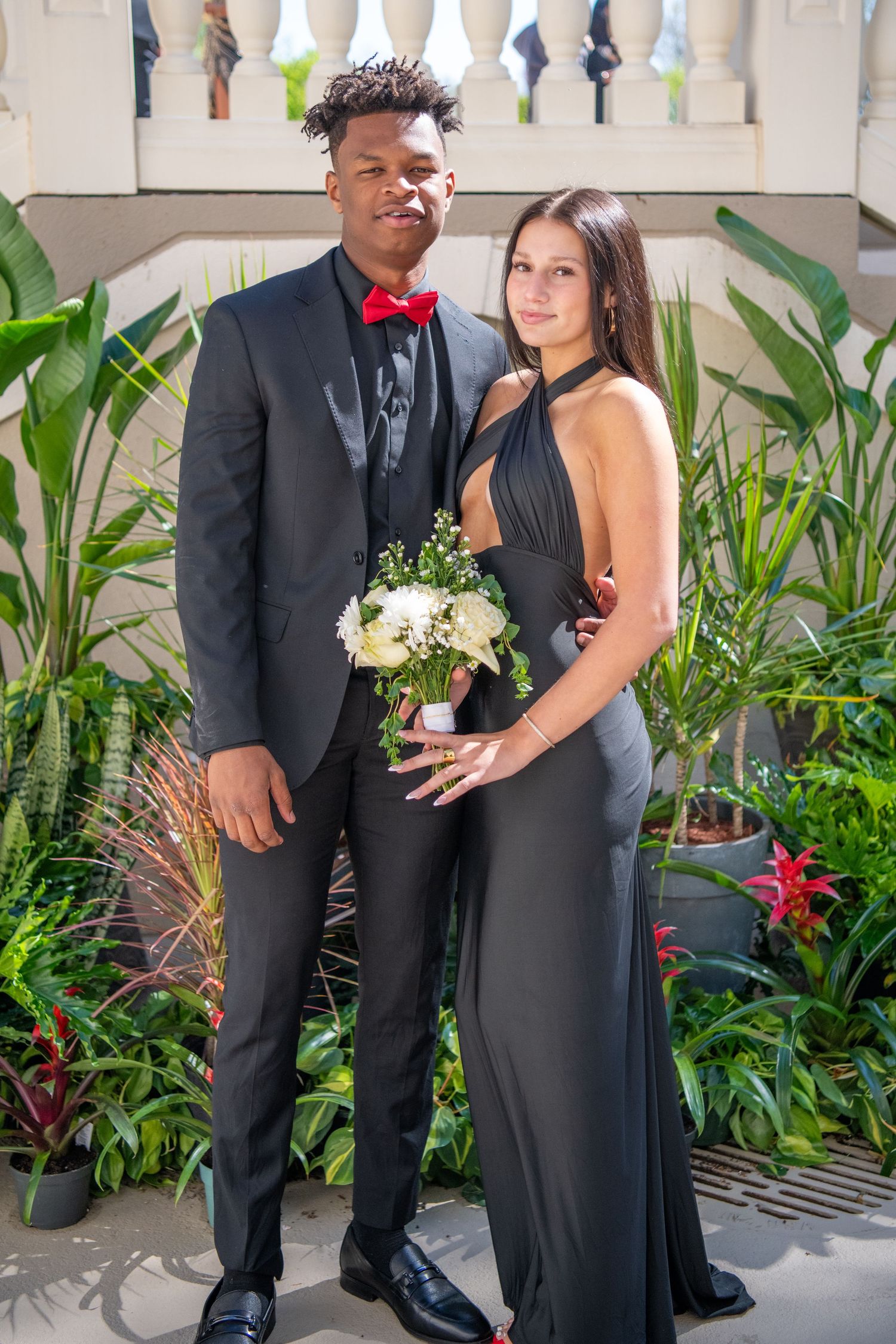 Formal couple standing in front of white railing and tropical plants at outdoor event.