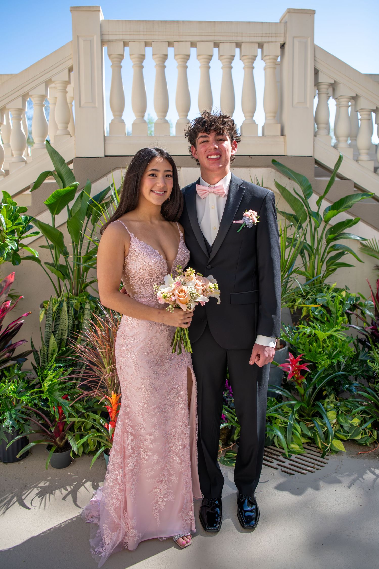Wedding couple poses against a white balustrade backdrop with tropical plants and flowers.