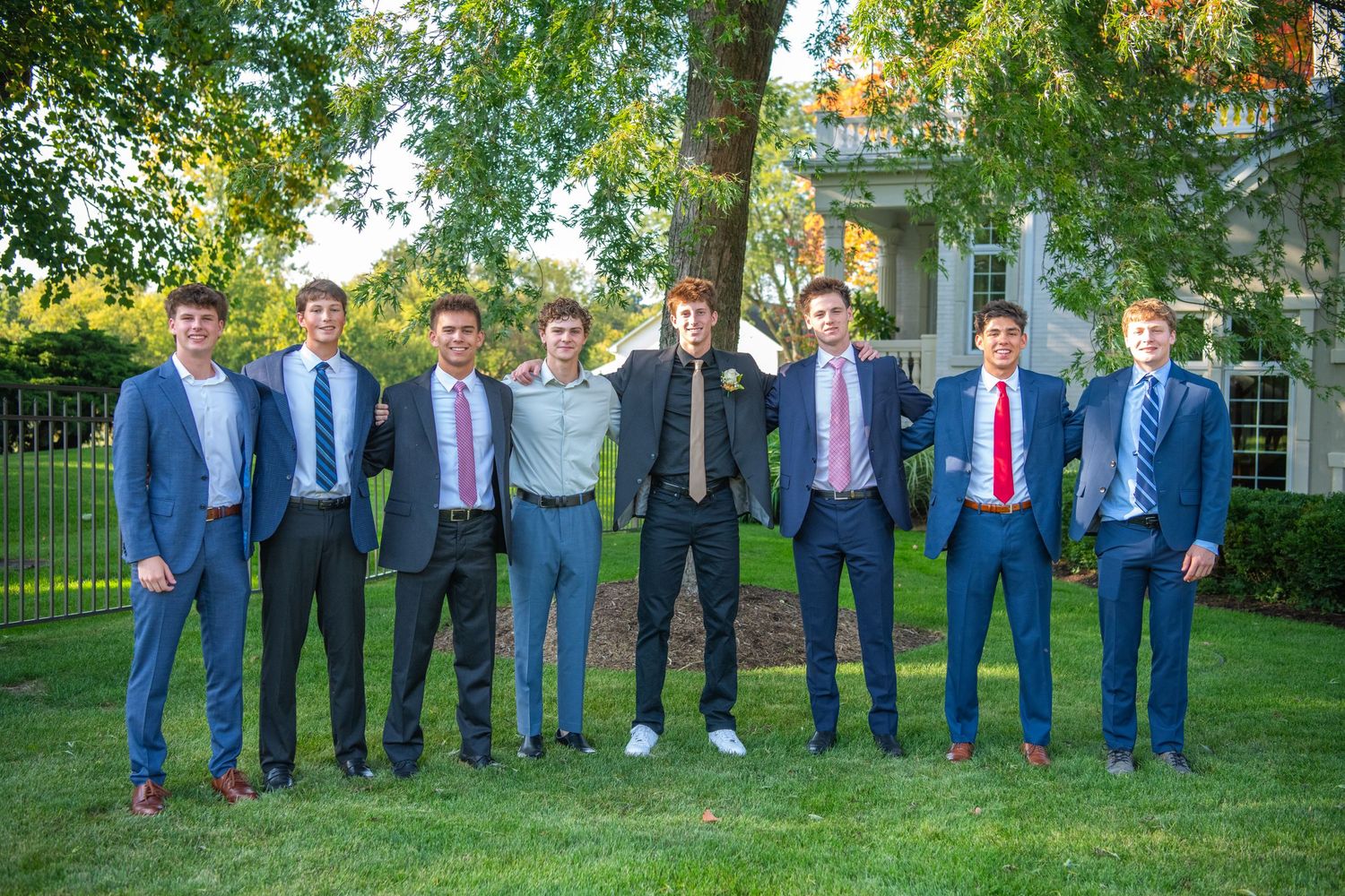 A group wearing blue suits and dress shirts poses together on a grassy lawn with trees in the background.