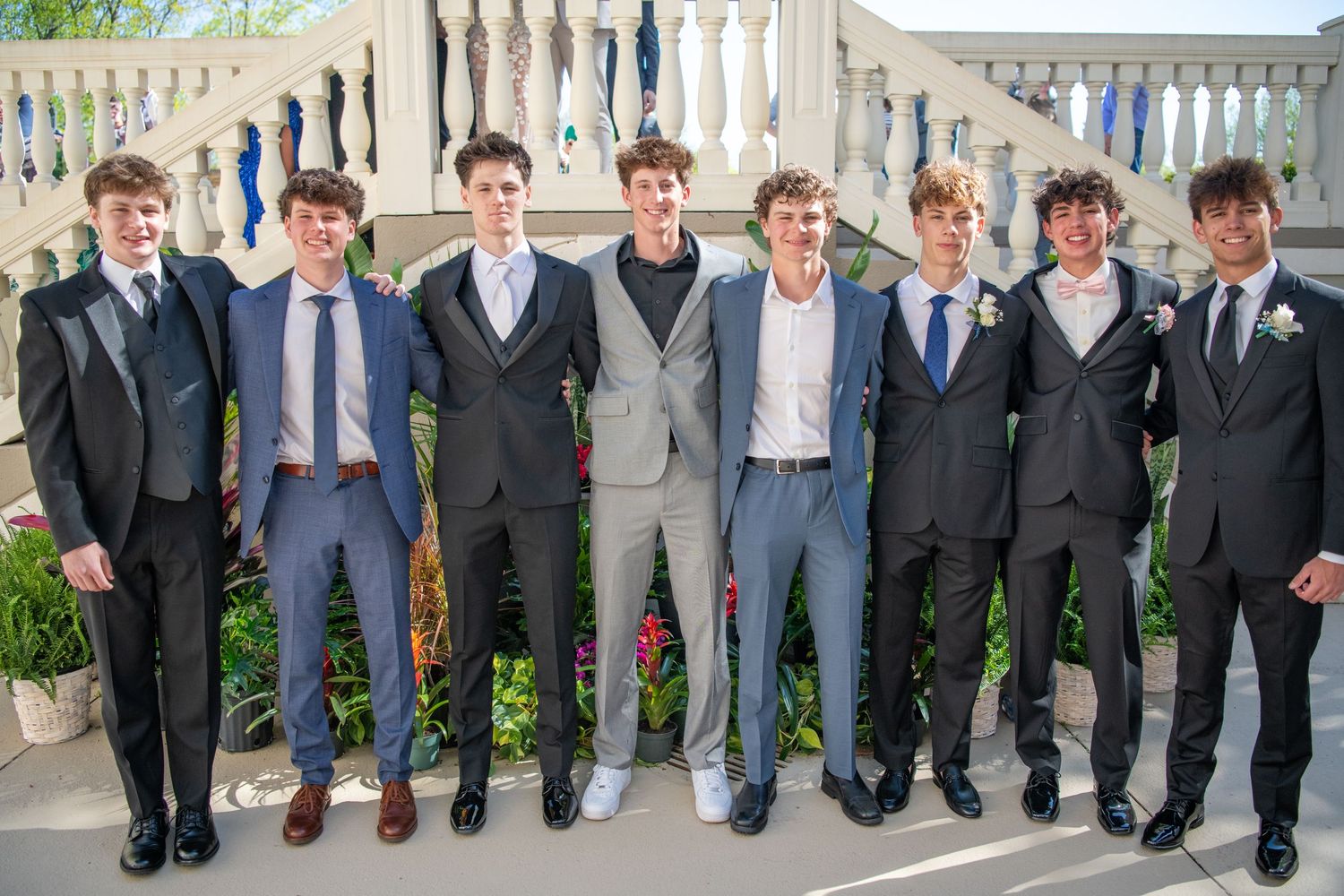 Row of groomsmen in suits stand against white balustrade showing their shoes in matching poses.