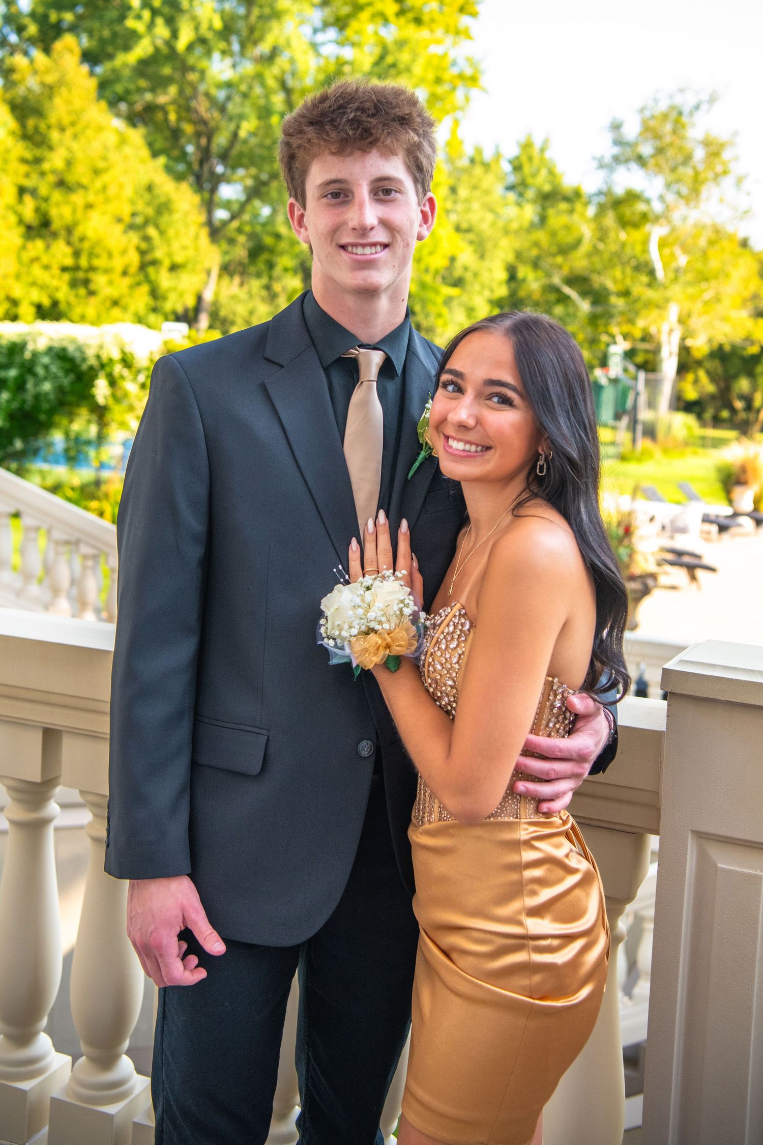 A couple dressed in formal attire poses together at a prom or formal event with yellow trees in the background.