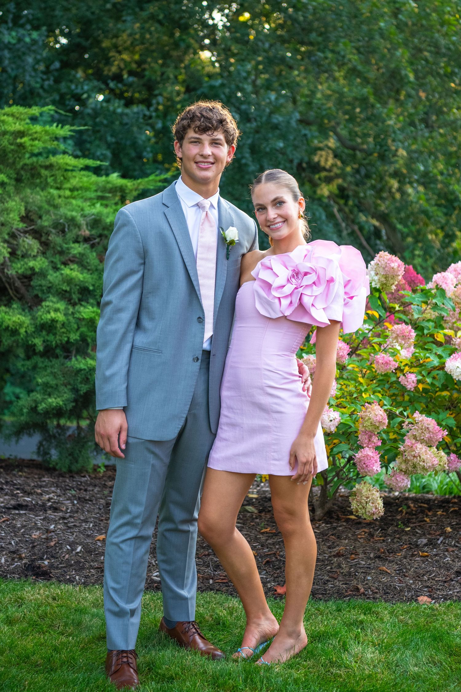 A couple poses together in a garden setting with blooming pink hydrangeas and greenery.