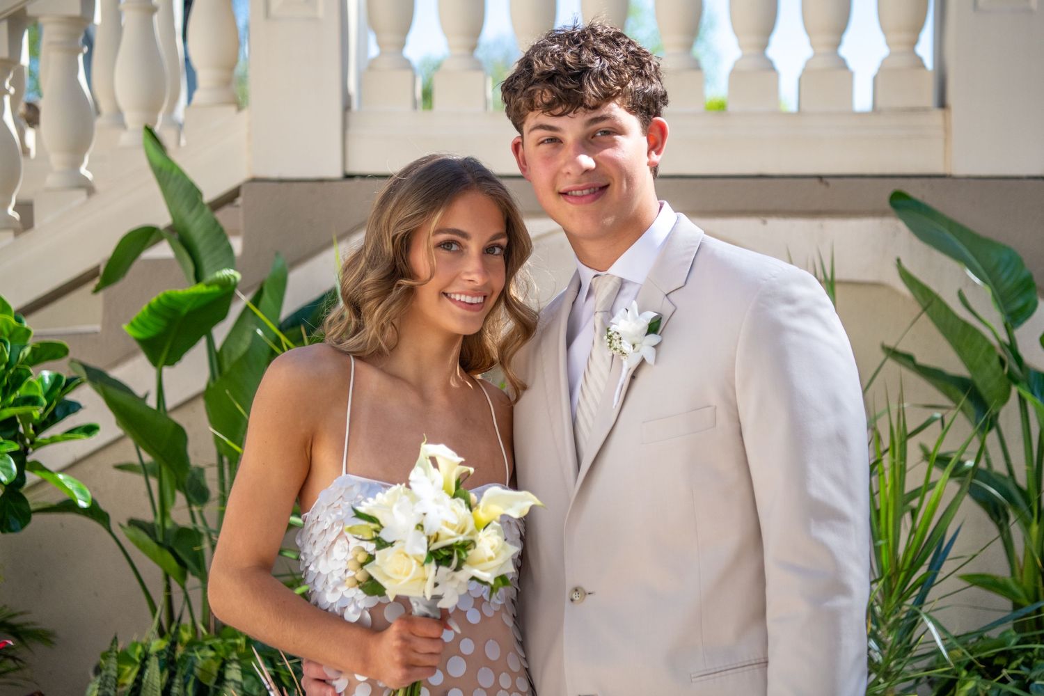 Elegantly dressed couple posing for photos against decorative white background with plants.