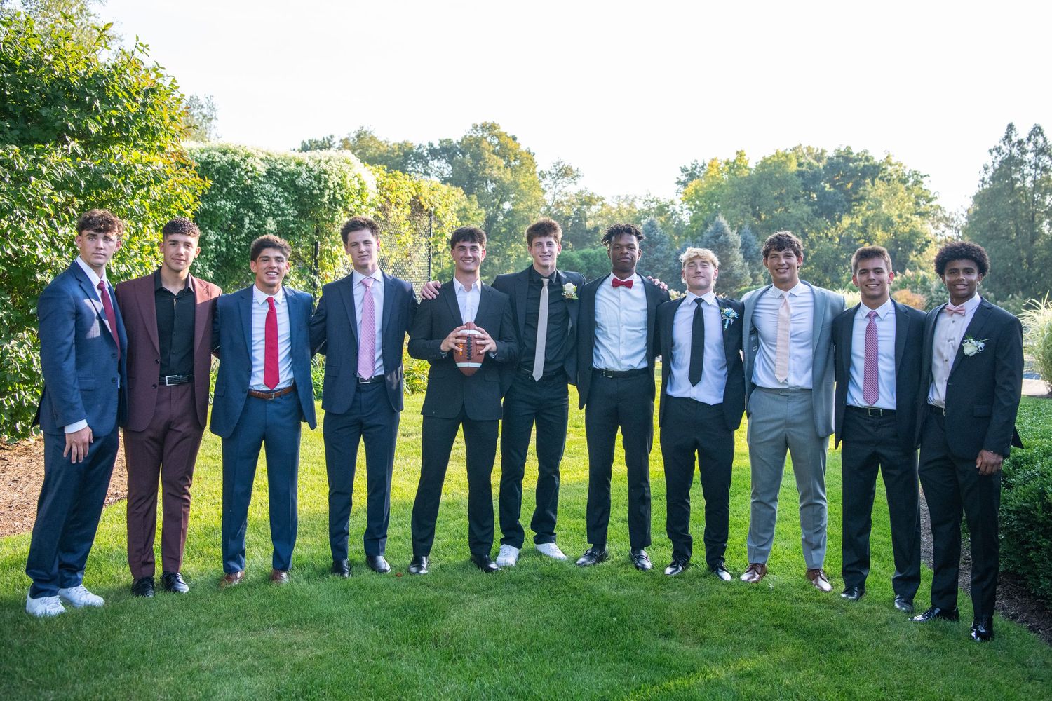 A group of people in formal attire stand in a line outdoors on a sunny day with trees in the background.