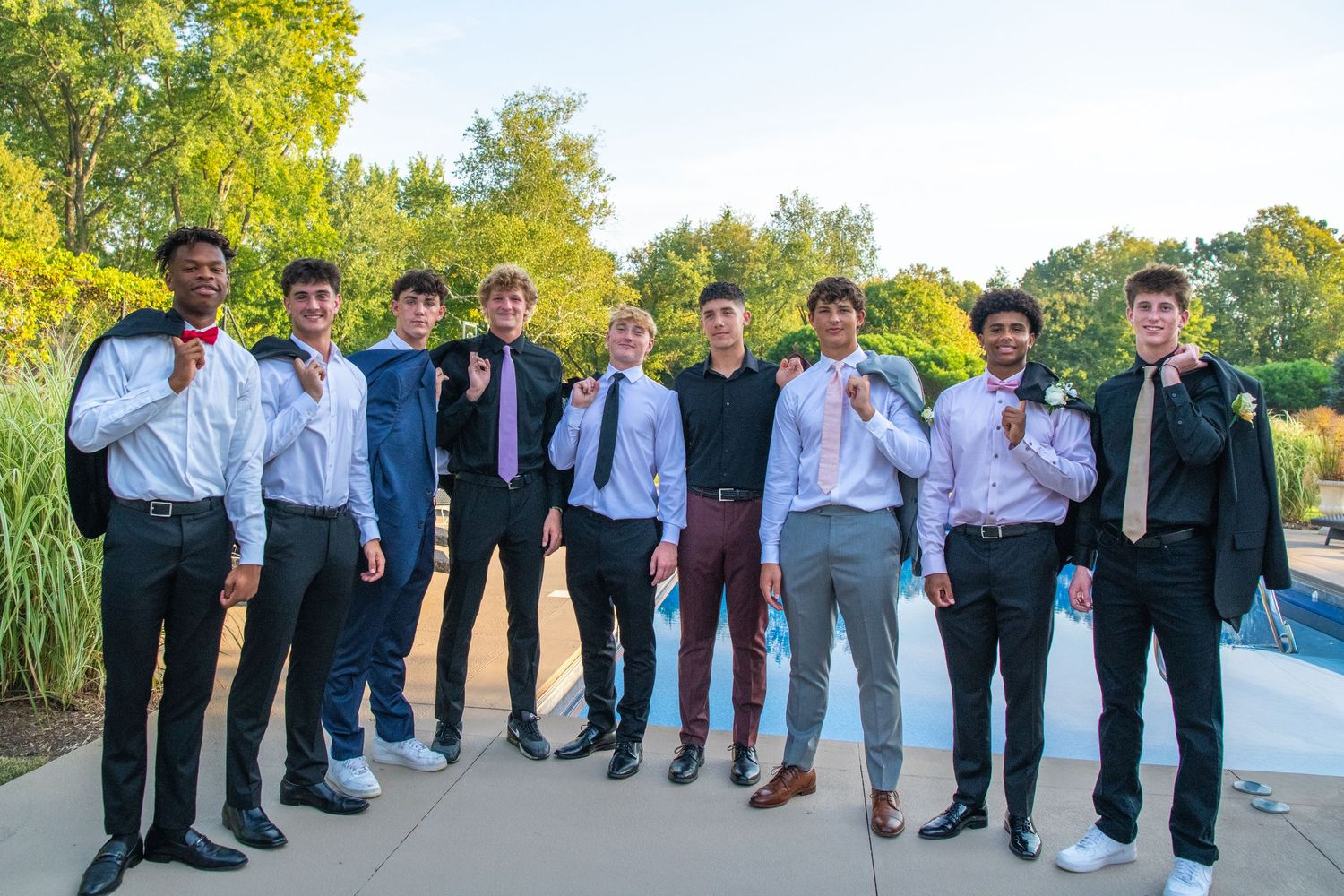 A group of formally dressed young people pose together outdoors against a natural backdrop with trees and greenery.