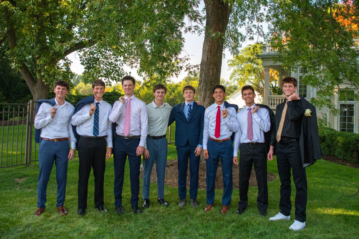 A line of people in formal attire stand together outdoors with green trees and foliage behind them.
