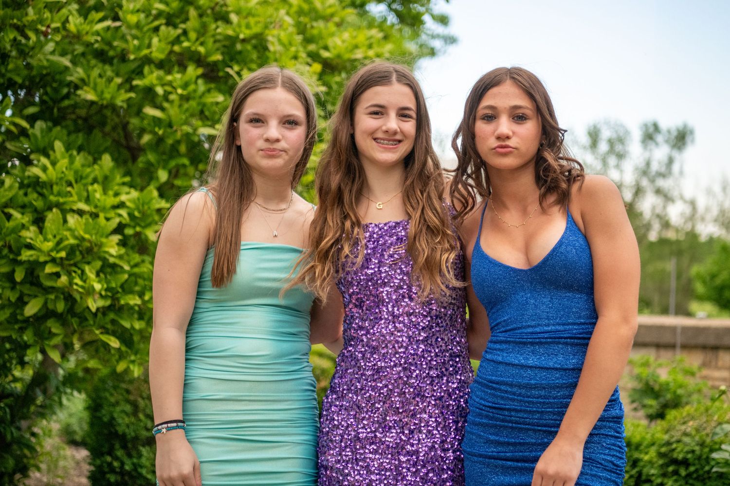 A trio poses in sequined rompers of different colors against a natural backdrop.