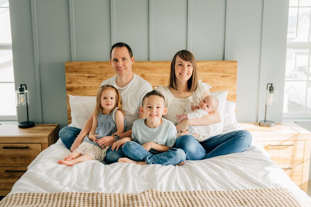 A happy family sits together on a bed with wooden headboard in a bright sunlit bedroom.