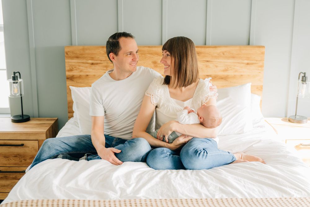 A couple sits together on a white bed with wooden headboard while gazing at each other lovingly.