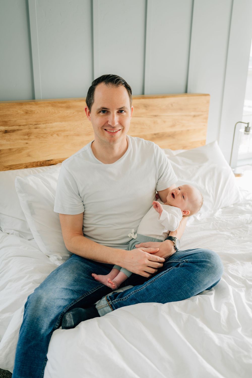 Someone sitting cross-legged on a white bed with a baby, wearing a white t-shirt and jeans against a wooden headboard.