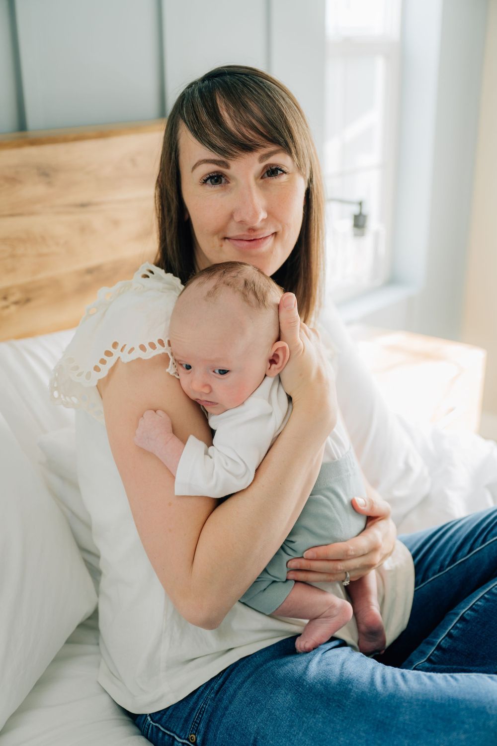 A mother and baby share an intimate moment while relaxing together on a bed near a bright window.