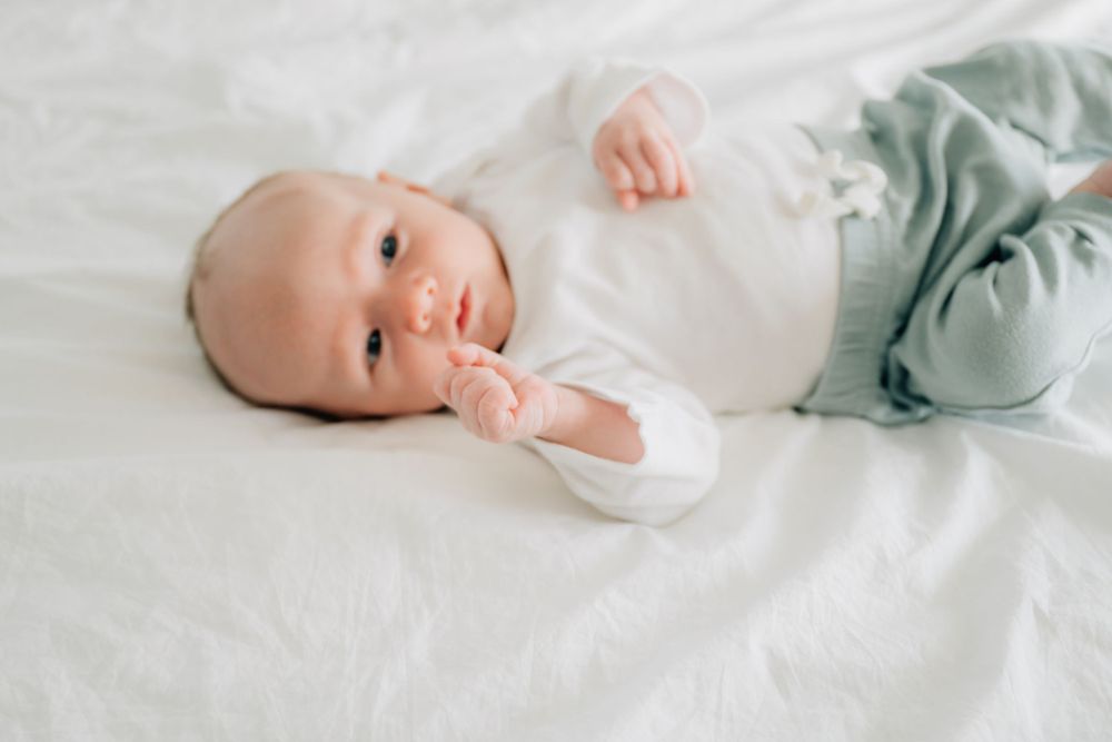 Sweet baby wearing white and mint green outfit laying on white bedding looking up at camera.