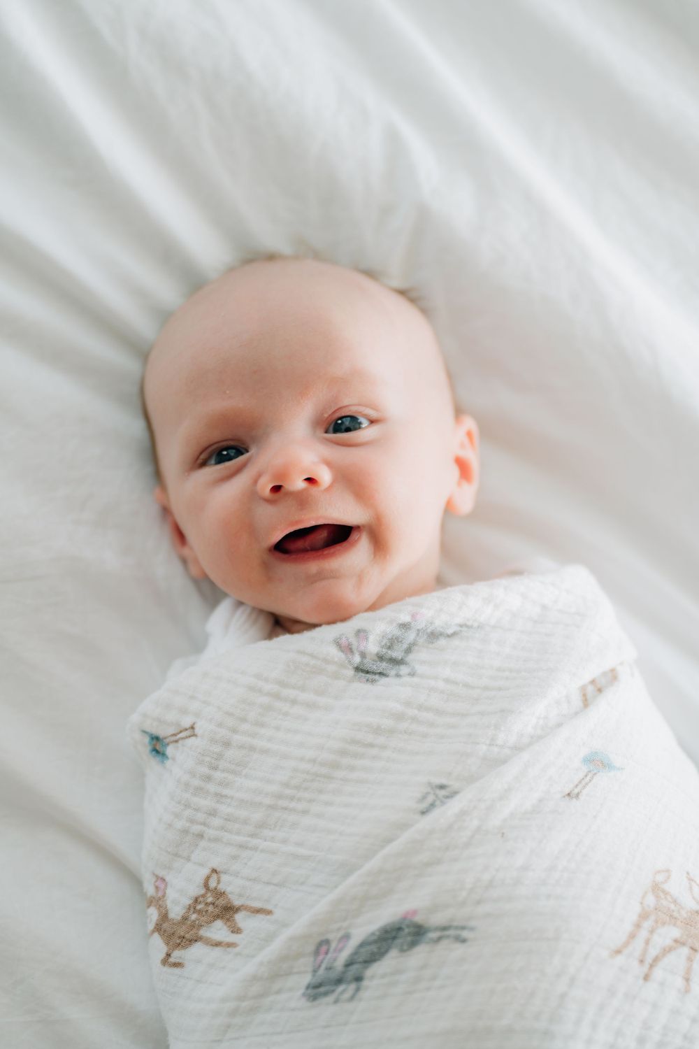A happy baby wrapped in a soft white muslin blanket with delicate animal prints smiles while lying down.