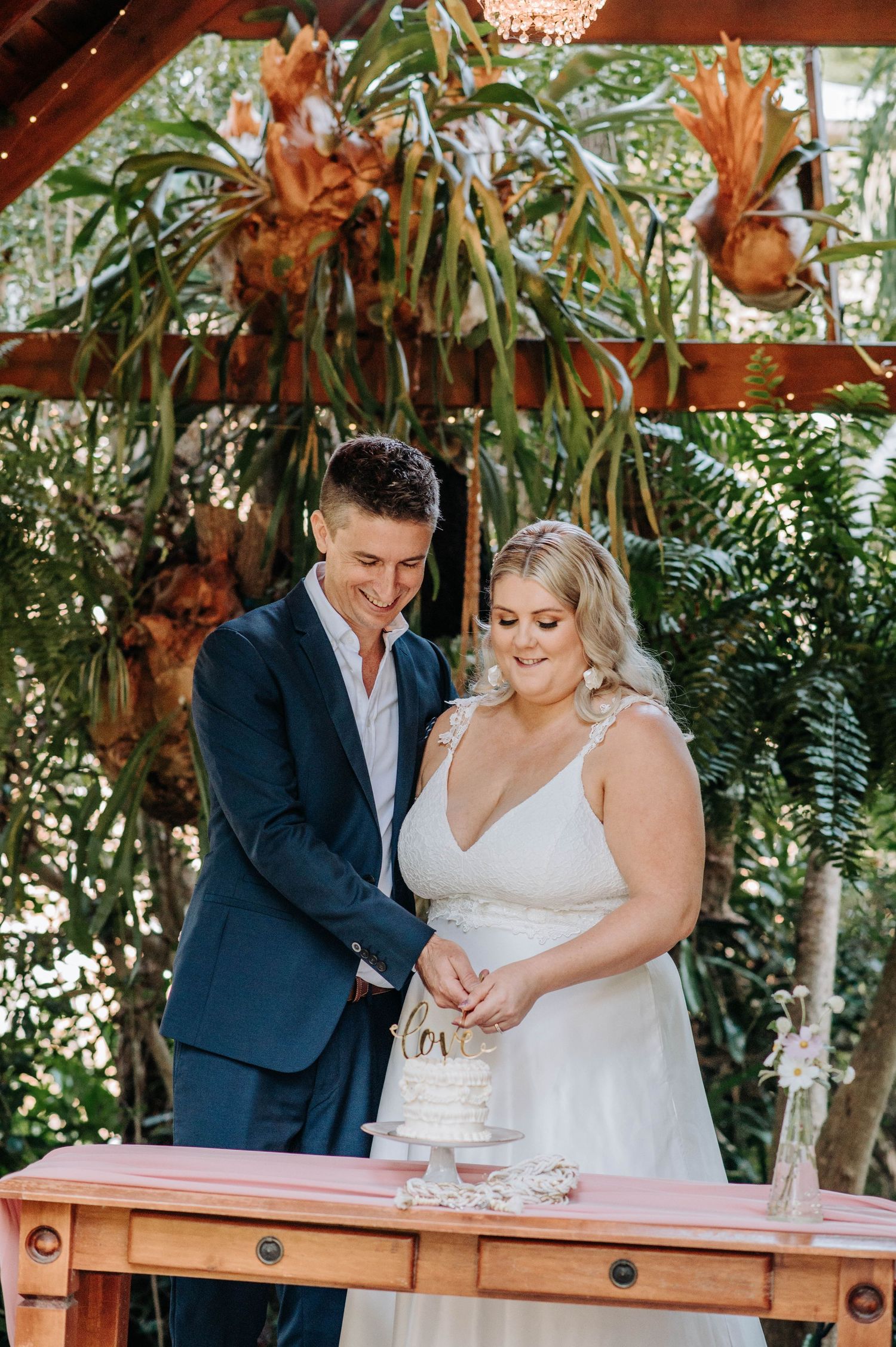 A couple sharing a joyful moment while cutting their wedding cake under decorative tropical foliage.