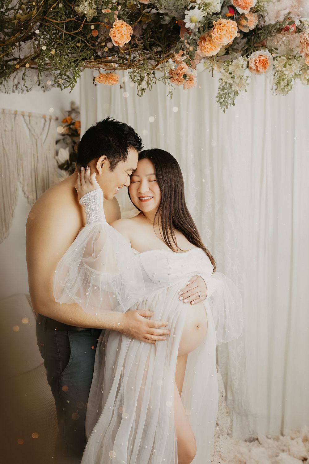 Romantic maternity photoshoot with couple under floral arch and white backdrop.