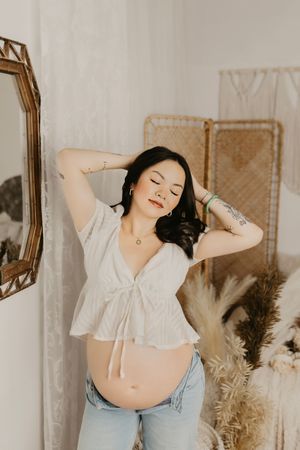 Casual maternity portrait with white top and jeans in a boho-styled room with mirror.