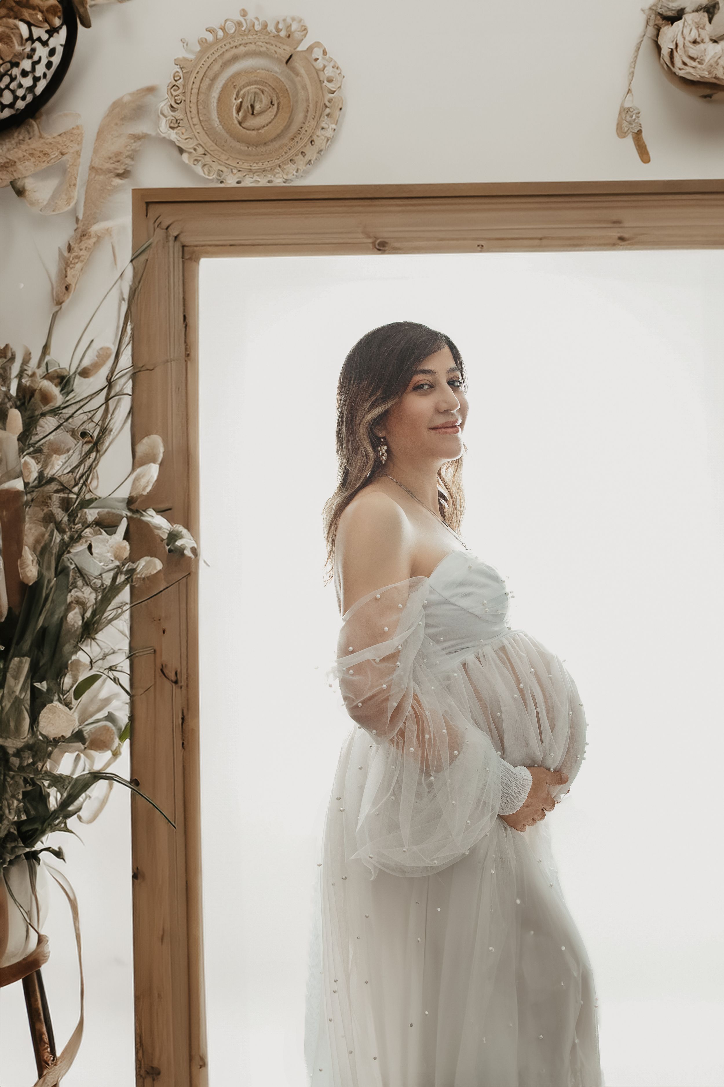 Ethereal maternity portrait in a white lace dress against a light backdrop with boho decorations.
