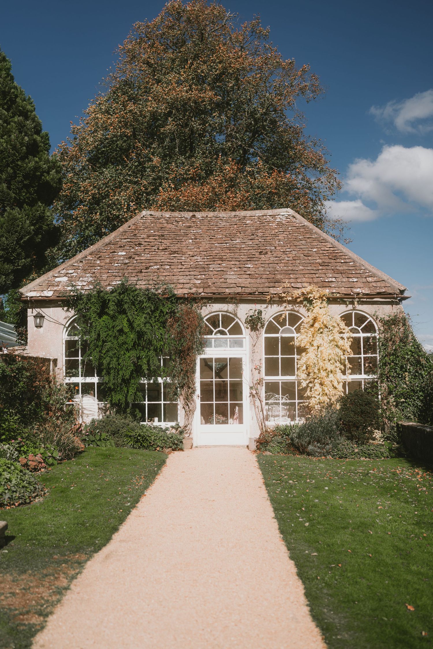 A charming white garden house with arched windows and a path leading to its entrance surrounded by landscaping.