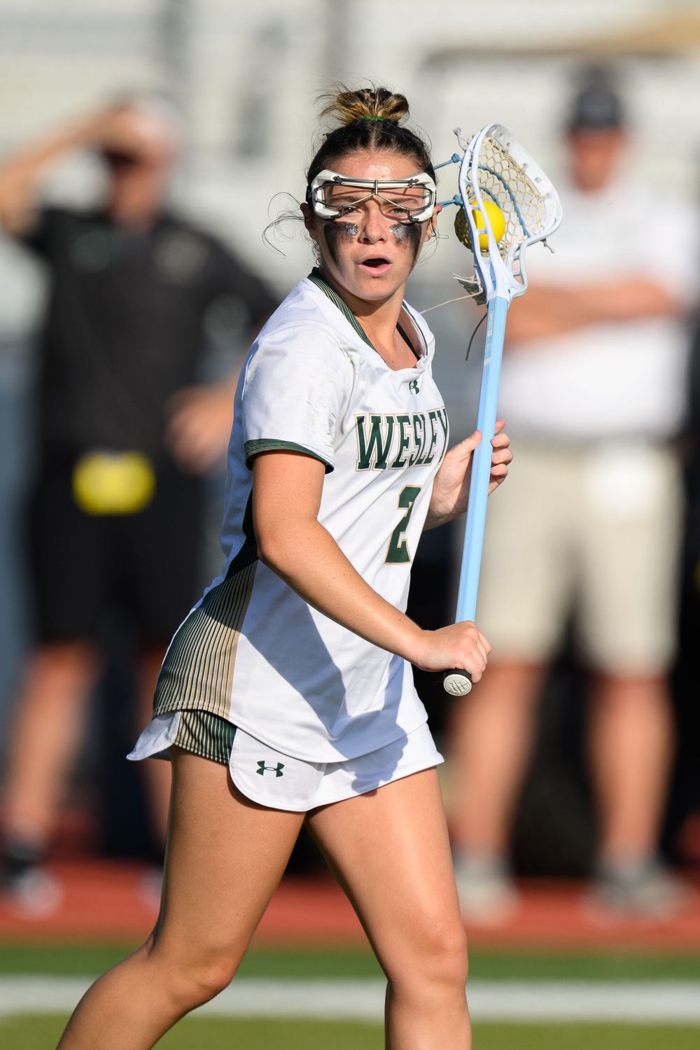 Women's lacrosse player in white uniform and protective eyewear cradles the ball during competitive match.