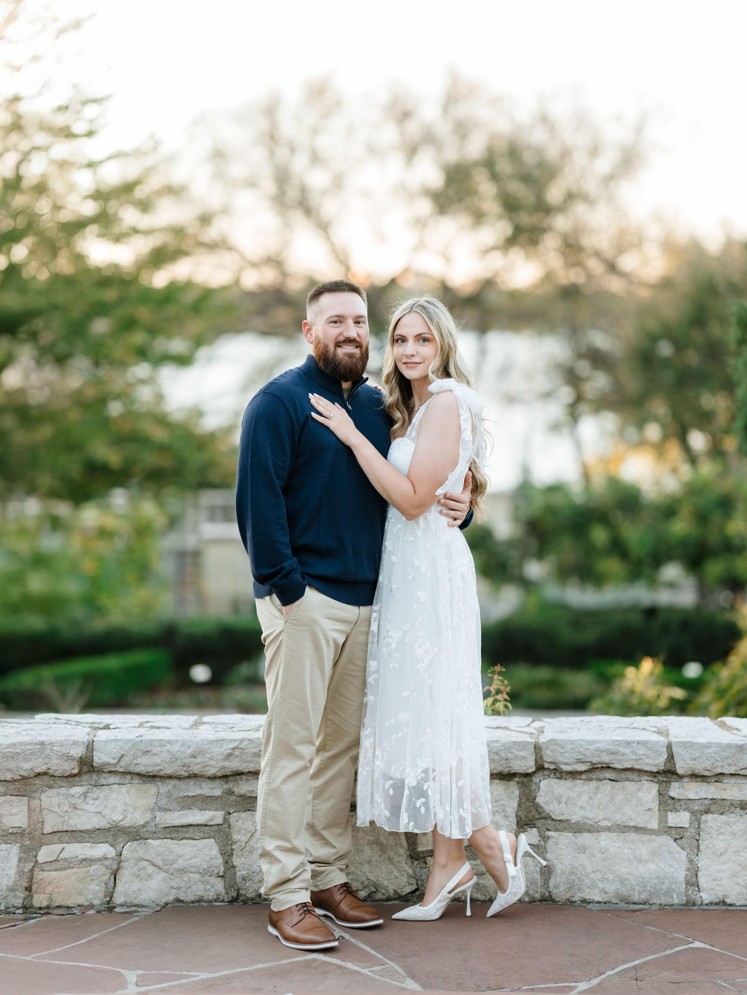 Romantic couple shares tender moments in white dress and navy shirt while standing on stone patio at sunset.
