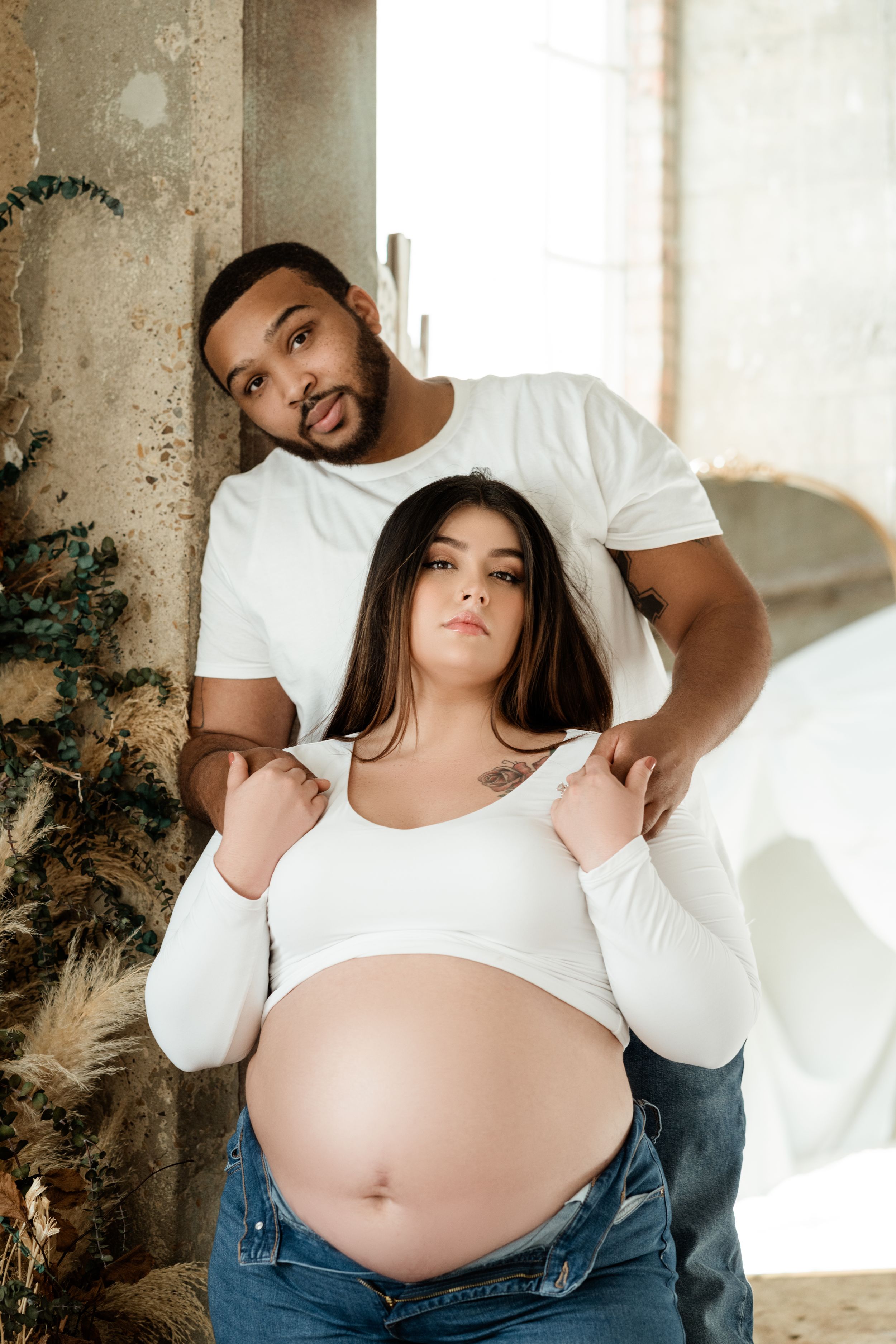 A couple poses for a maternity photo, with the pregnant woman in white and jeans standing against a rustic wall in natural light.