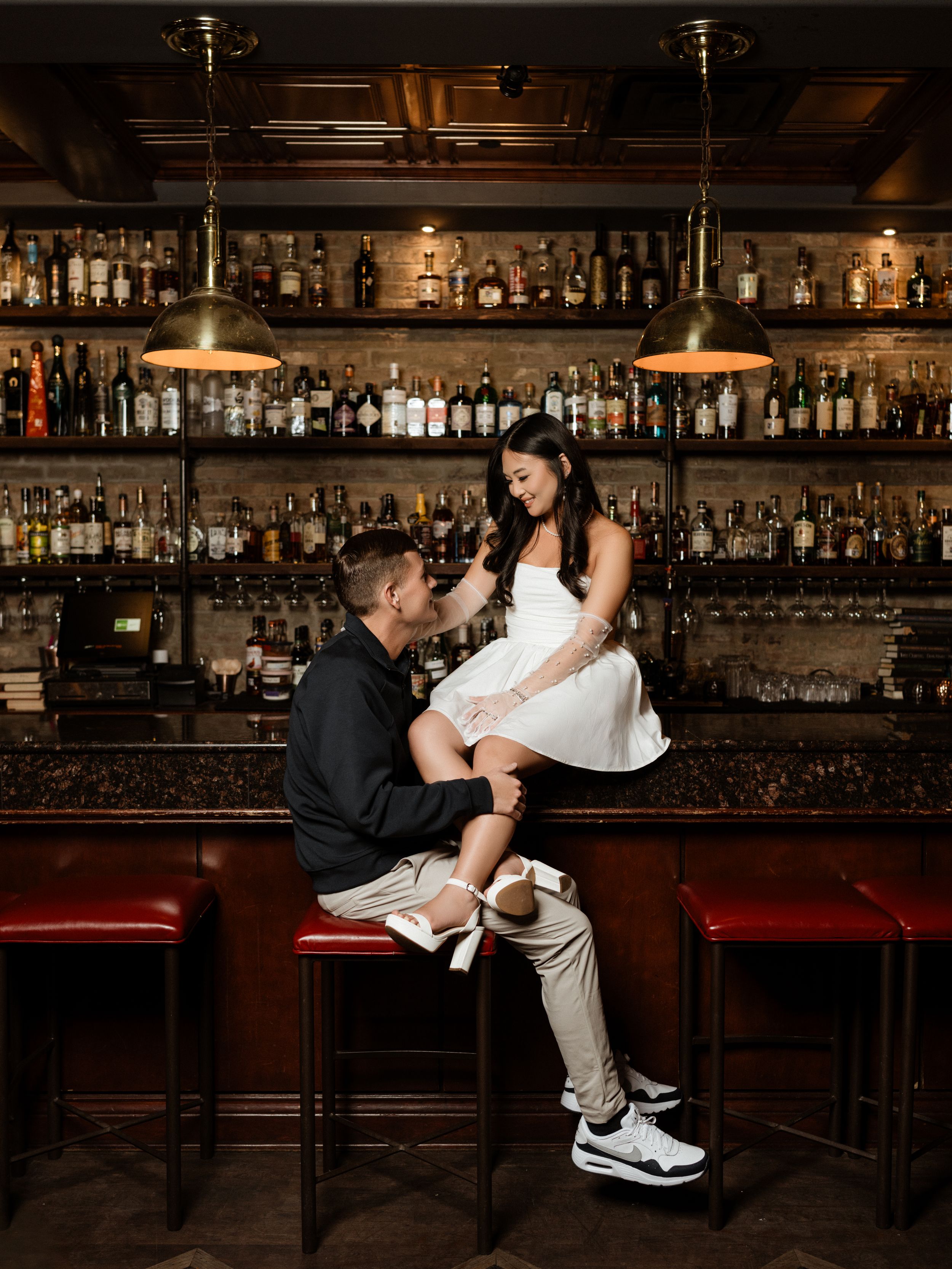 Cozy bar interior with vintage wooden shelving displaying numerous liquor bottles illuminated by pendant lights.