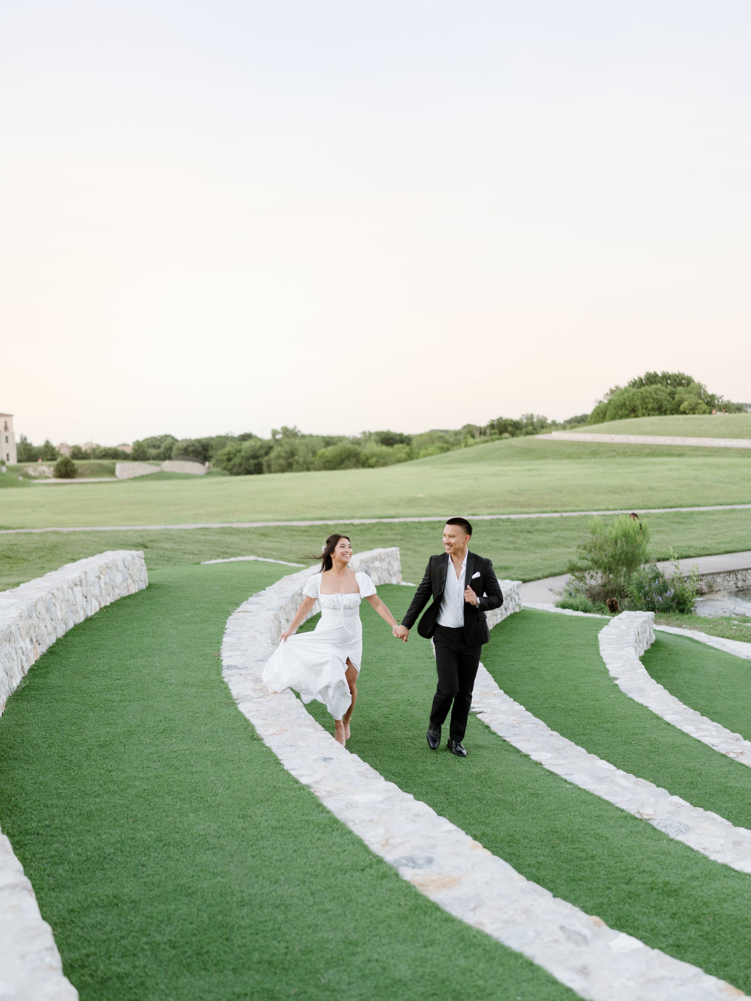 Wedding couple runs playfully along curved stone pathways on a manicured lawn.
