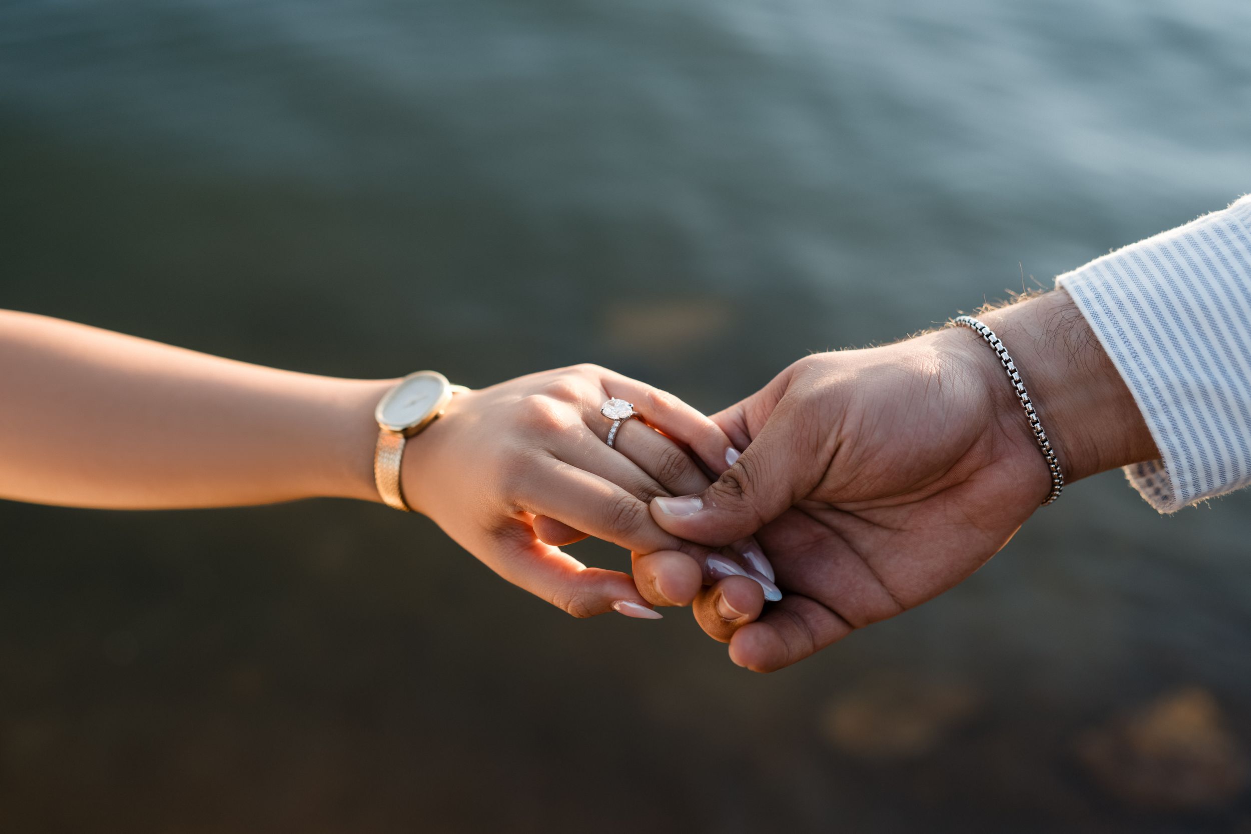 Two hands tenderly clasping together in a romantic moment with water in the background during golden hour.