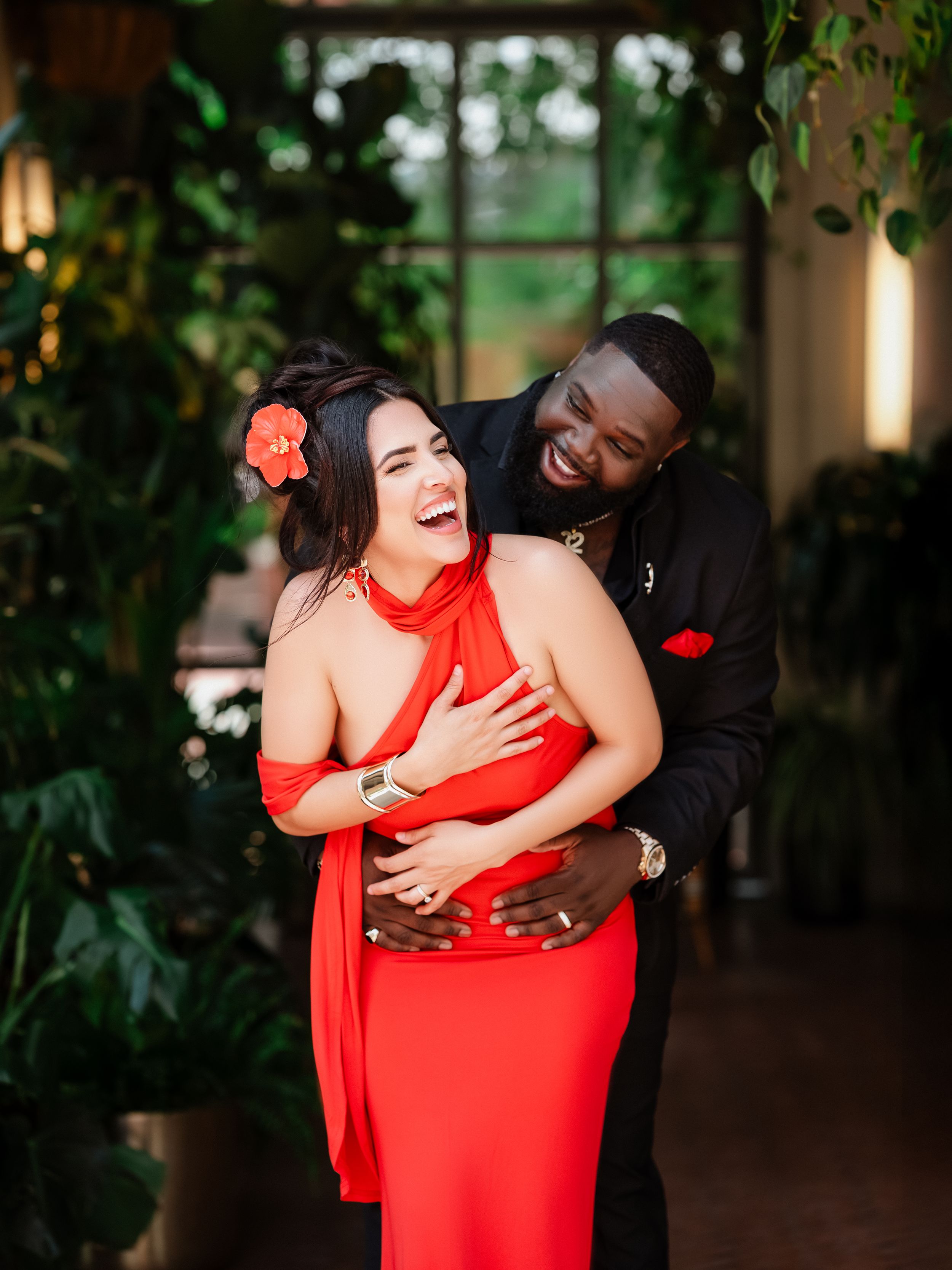 Joyful couple in red dress and black suit share intimate embraces and laughter in a romantic garden setting.