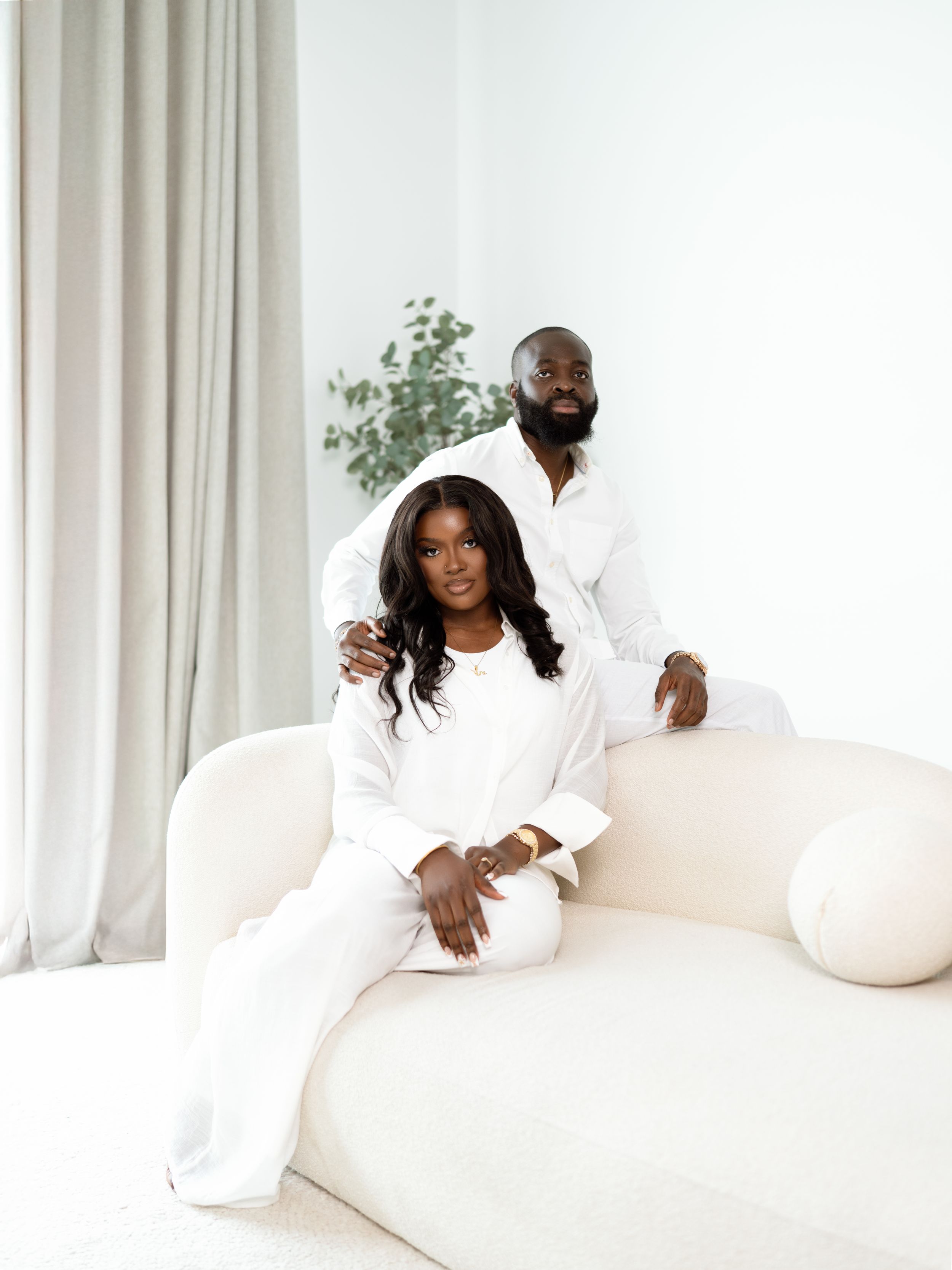 A picture of two people dressed in white sitting together on a cream-colored couch in a minimalist white space.