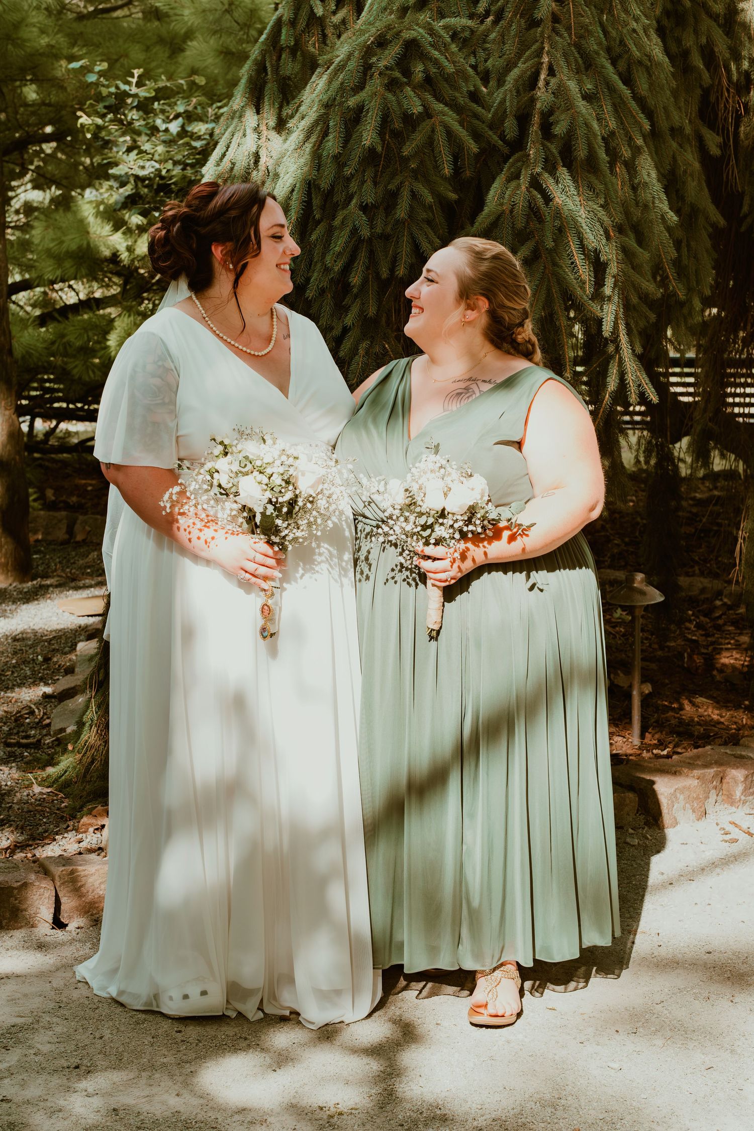 Two women in flowing dresses hold bouquets while sharing a laugh in a garden setting.