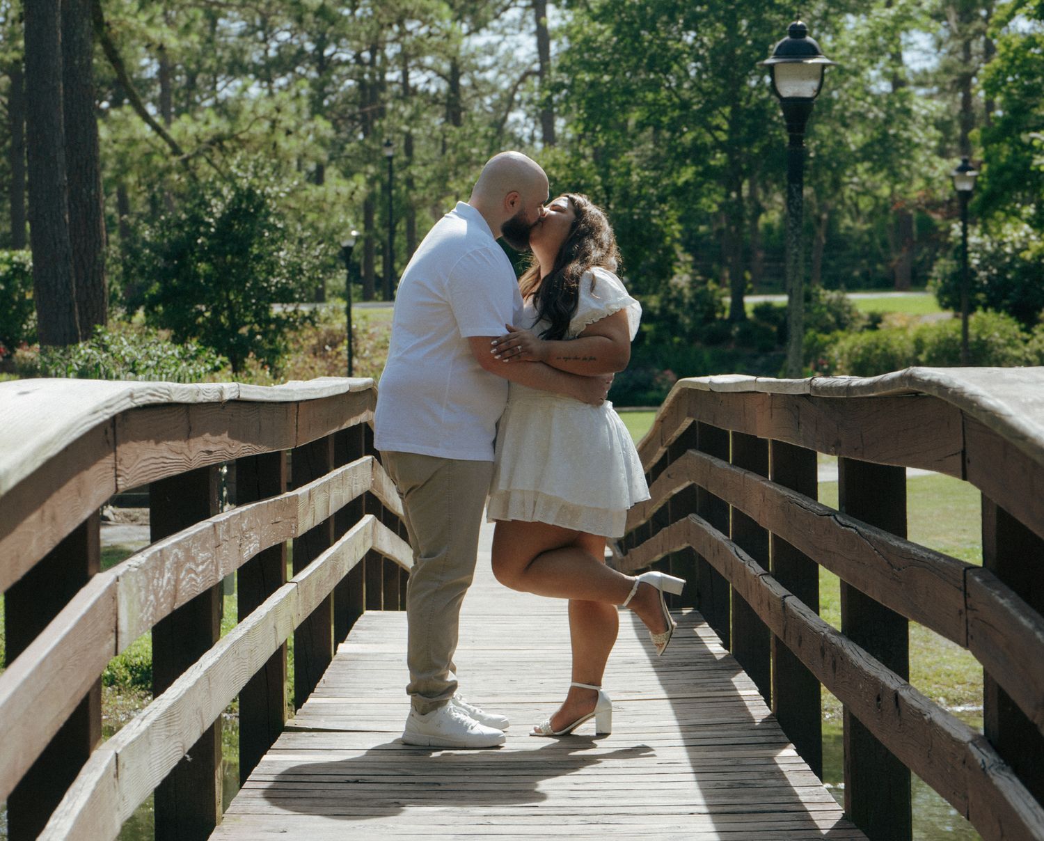 A couple embraces while standing on a wooden bridge surrounded by trees in a park setting.