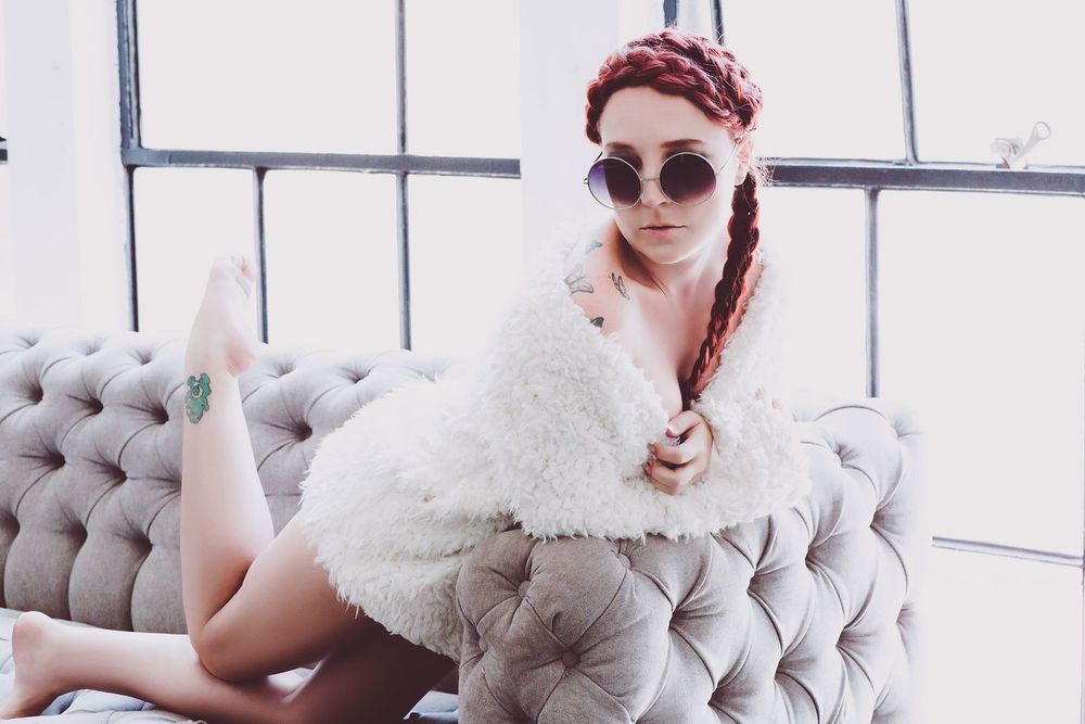 Person in sunglasses lounges on a grey tufted couch wearing a fluffy white sweater and red headscarf.