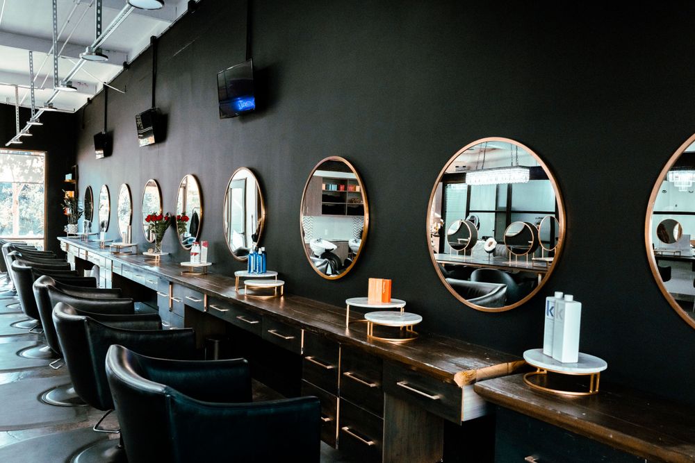 Modern hair salon interior with round mirrors mounted on dark walls and blue styling chairs arranged in a row.