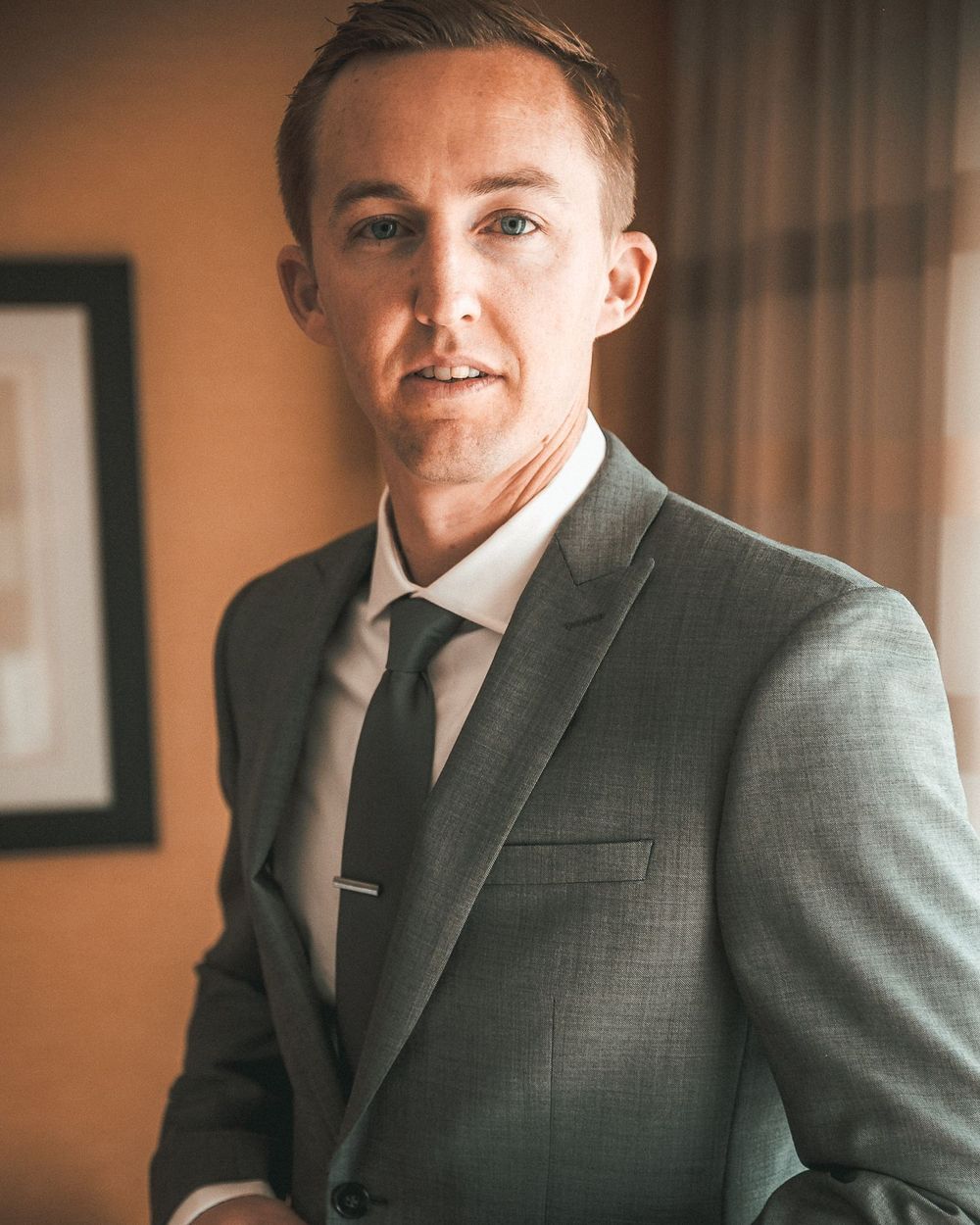 Professional portrait of someone wearing a grey suit and tie against a warm-toned background with framed artwork.