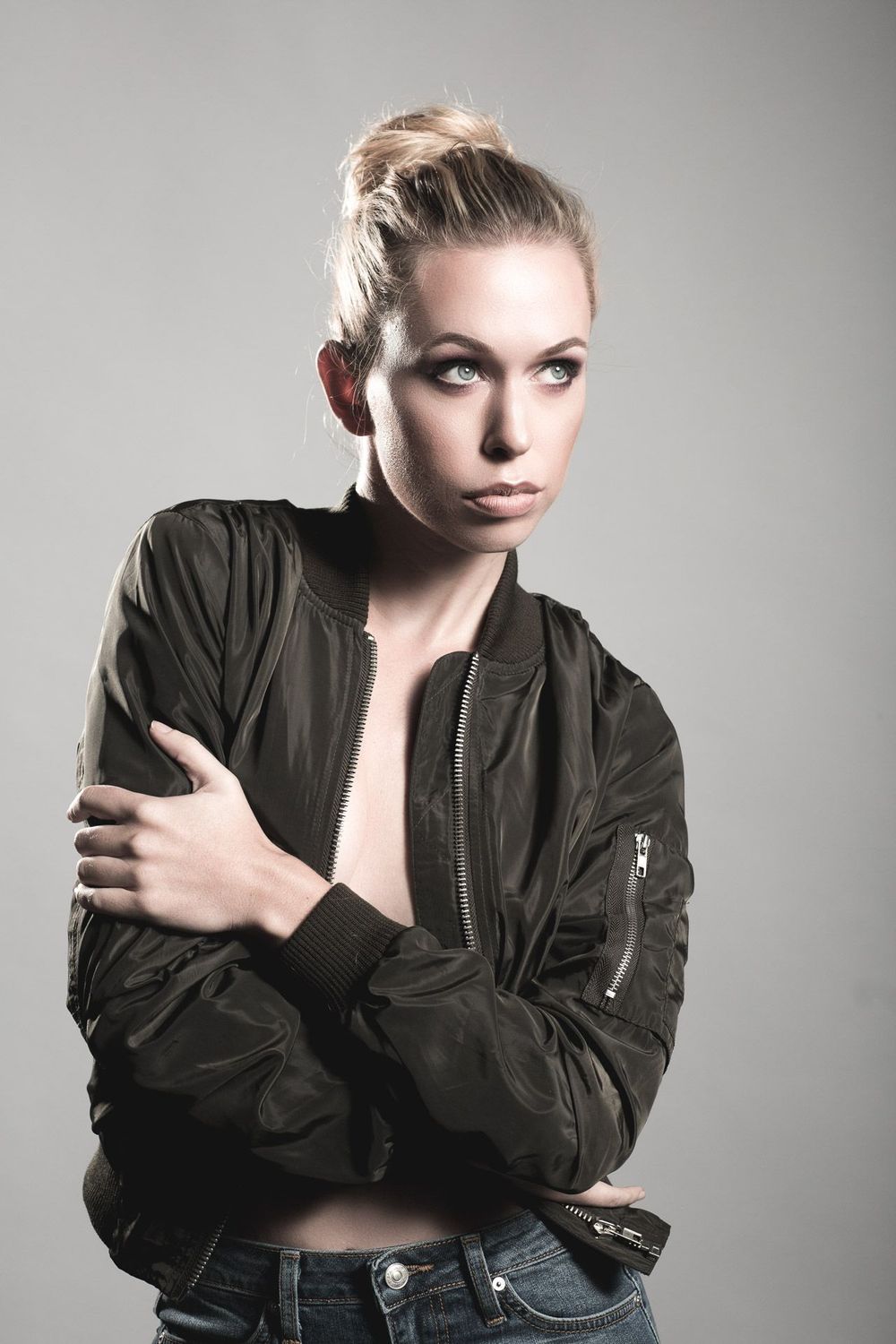 Fashion portrait with olive bomber jacket and blonde hair bun against gray background.