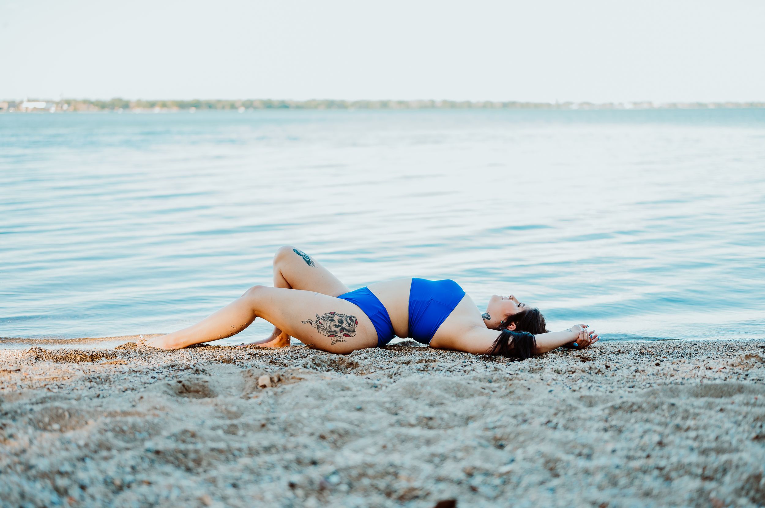 Woman in blue swimsuit poses artistically on beach by calm water.